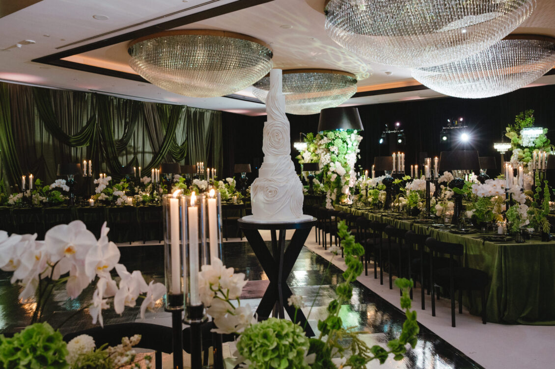 Elegant event hall with green drapery, floral centerpieces, tall candelabras, and a white sculpted cake on a pedestal. Large chandeliers hang from the ceiling, creating a luxurious atmosphere—perfect for a black-tie wedding at Hotel X Toronto.