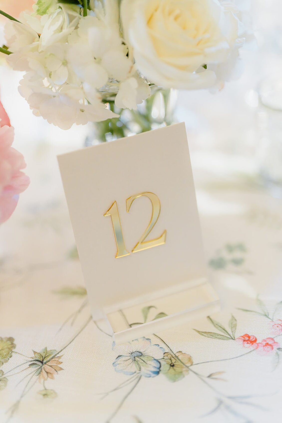 A white card with the number 12 in gold stands on a floral tablecloth, surrounded by white and cream flowers in soft focus—capturing the elegance of an Irish castle wedding at Ashford Castle.