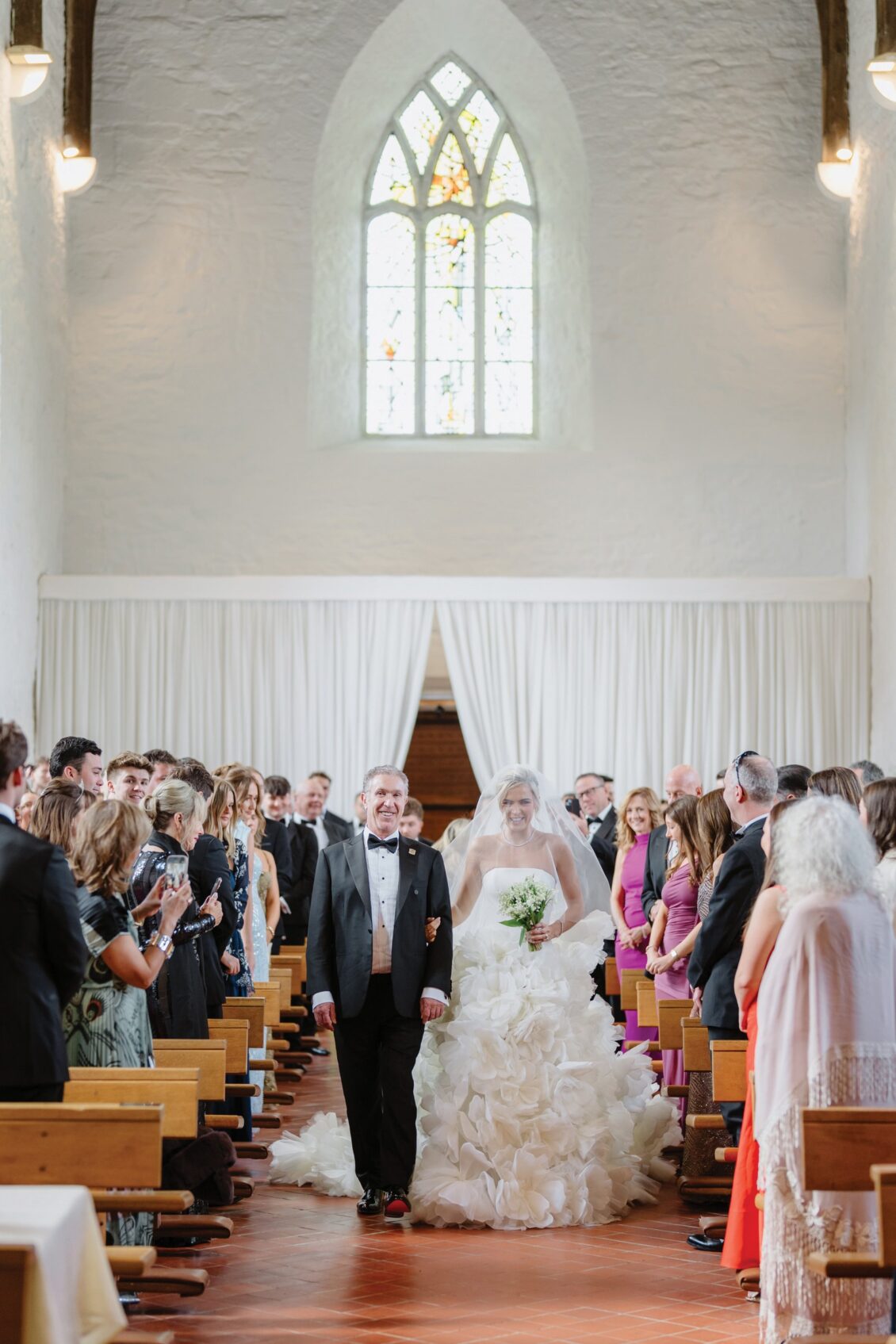 A bride in a white gown walks down the aisle with an older man in a tuxedo, surrounded by smiling wedding guests in the sunlit halls of Ashford Castle, where wooden benches and a large arched window evoke the timeless elegance of an Irish castle wedding.