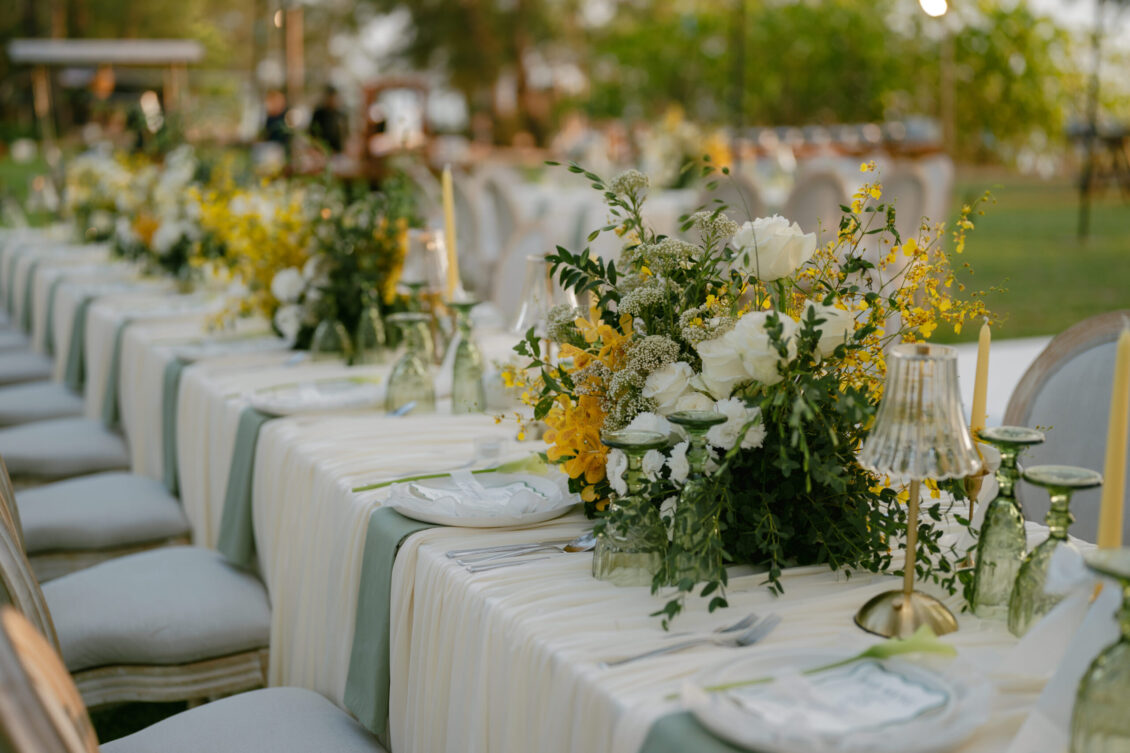 Elegant outdoor dining table set with white linens, green runners, and large floral arrangements with white and yellow flowers; glassware, plates, and candles arranged neatly—perfect for a Renaissance Phuket wedding. Lush greenery in the background completes the picturesque setting.