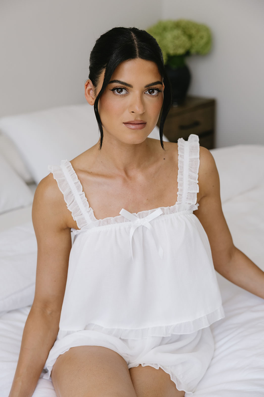 Erica, a woman with dark hair in a white ruffled top, sits on a bed with white bedding, looking directly at the camera. A plant with green flowers is visible on a bedside table in the background.