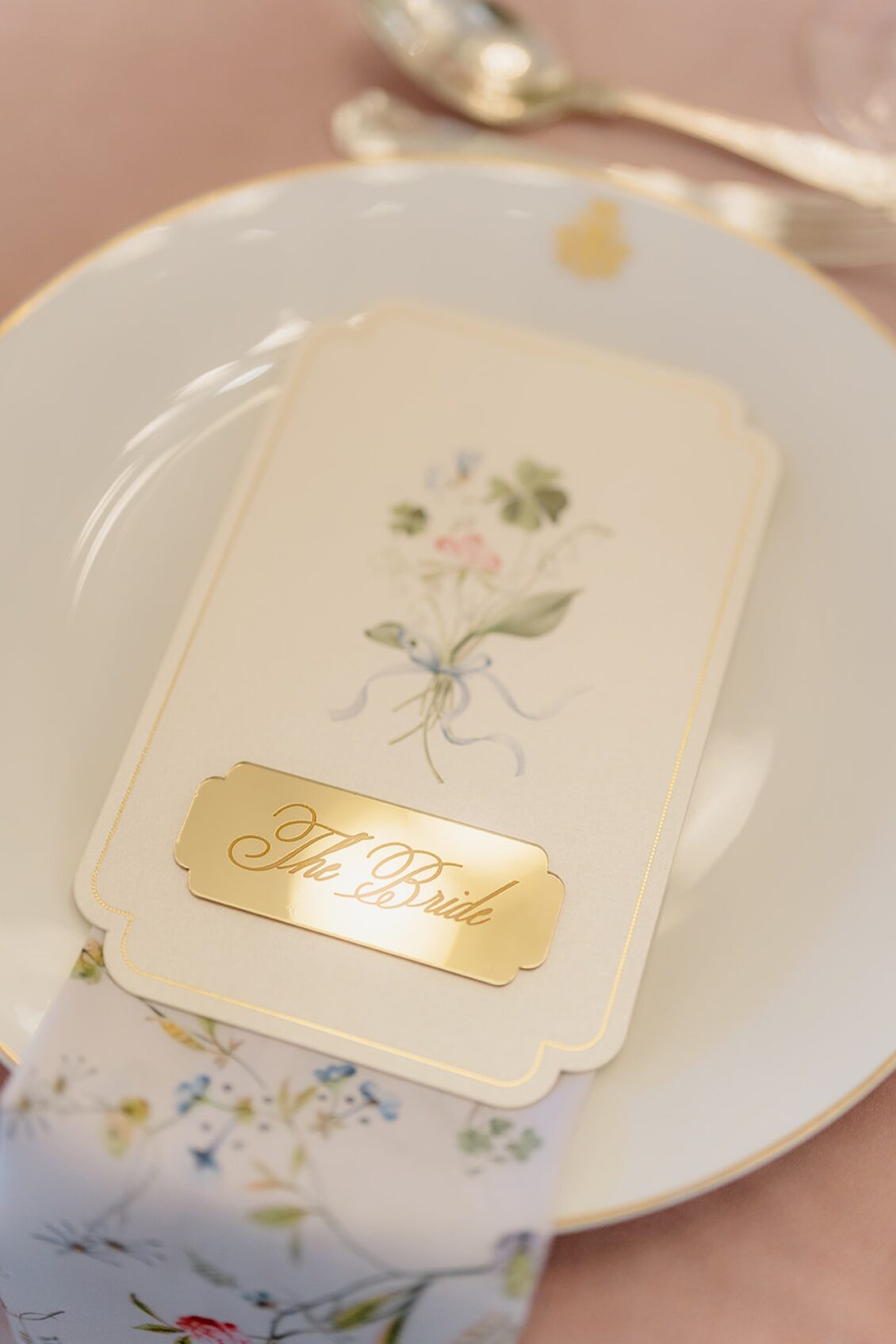 A white plate with a floral card labeled “The Bride” in gold script, resting atop a floral-patterned napkin, with a silver spoon and fork nearby on a pink tablecloth—setting the scene for an elegant Irish castle wedding at Ashford Castle.