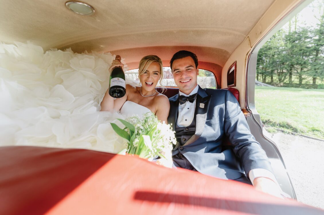 A bride and groom in wedding attire sit in the back of a vintage car after their Irish castle wedding at Ashford Castle. The bride holds a bouquet and a champagne bottle, both smiling excitedly as sunlight streams through the window onto their happy faces.