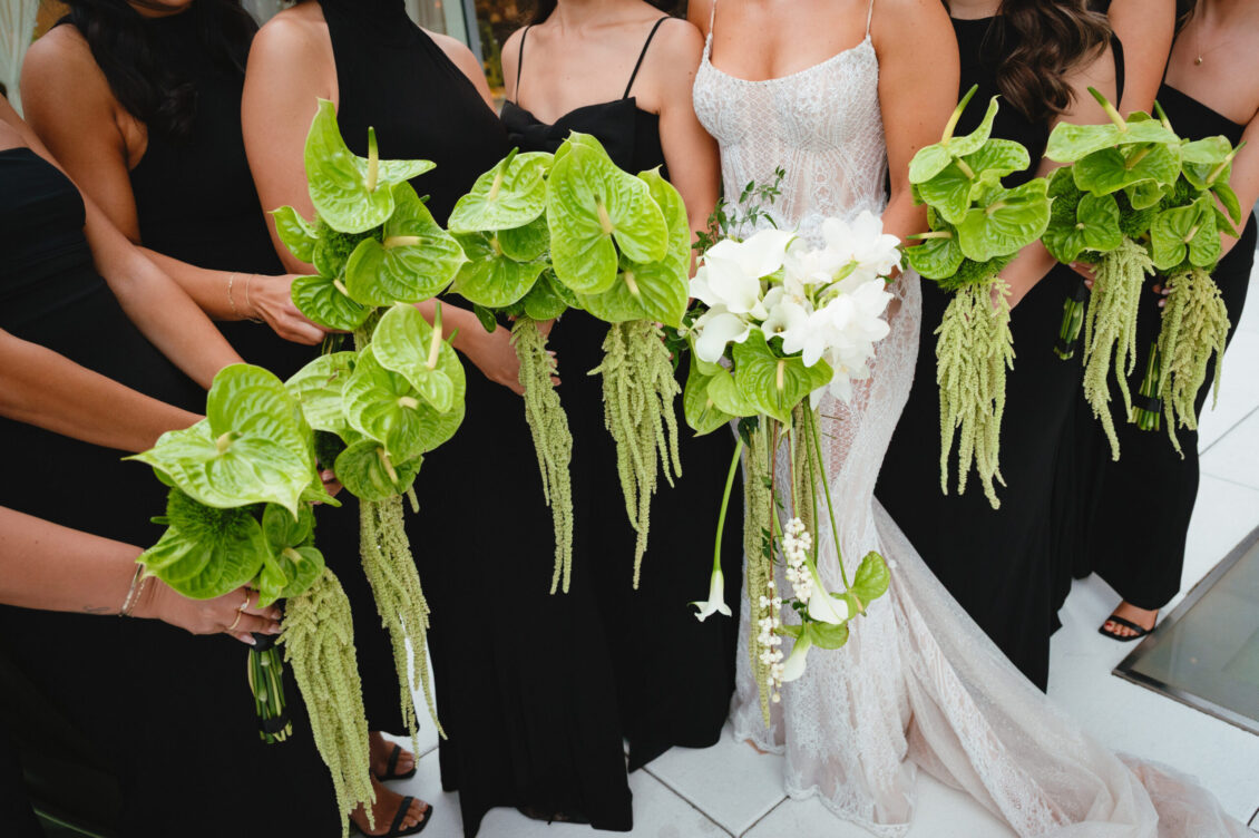 A bride in a white gown and bridesmaids in black dresses hold unique green tropical bouquets with long hanging foliage, perfectly complementing the sophisticated ambiance of a black-tie wedding at Hotel X Toronto.