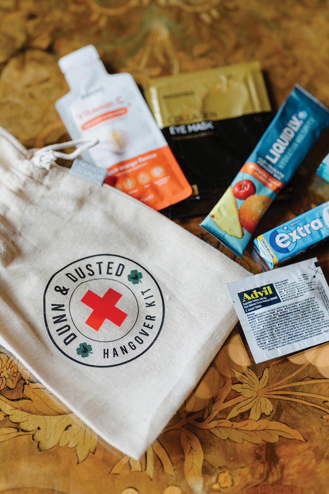 A canvas bag labeled “Down & Dusted Hangover Kit” on a table, surrounded by Advil, Extra gum, Liquid I.V., an eye mask, and a hydration packet.