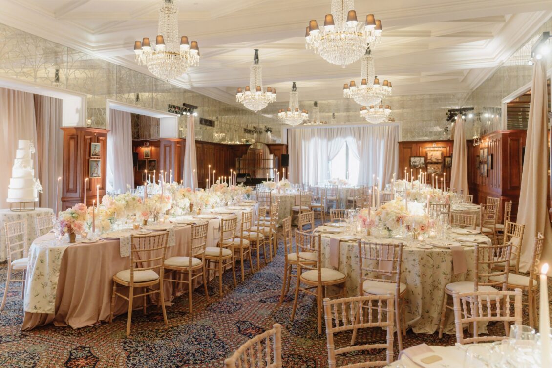 Elegant banquet hall with chandeliers, round tables with floral centerpieces, candles, and wooden chairs. The room evokes the timeless romance of an Irish castle wedding at Ashford Castle, set for a formal event with a wedding cake in the corner and soft, warm lighting throughout.