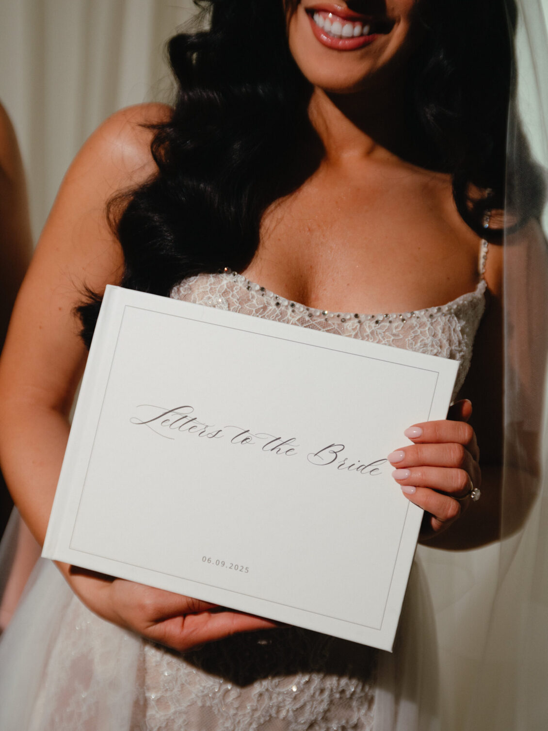 A bride in a lace wedding dress holds a white book titled “Letters to the Bride,” with the date 06.09.2025 printed at the bottom, capturing an elegant moment from her black-tie wedding at Hotel X Toronto. She smiles, partially visible from the shoulders up.