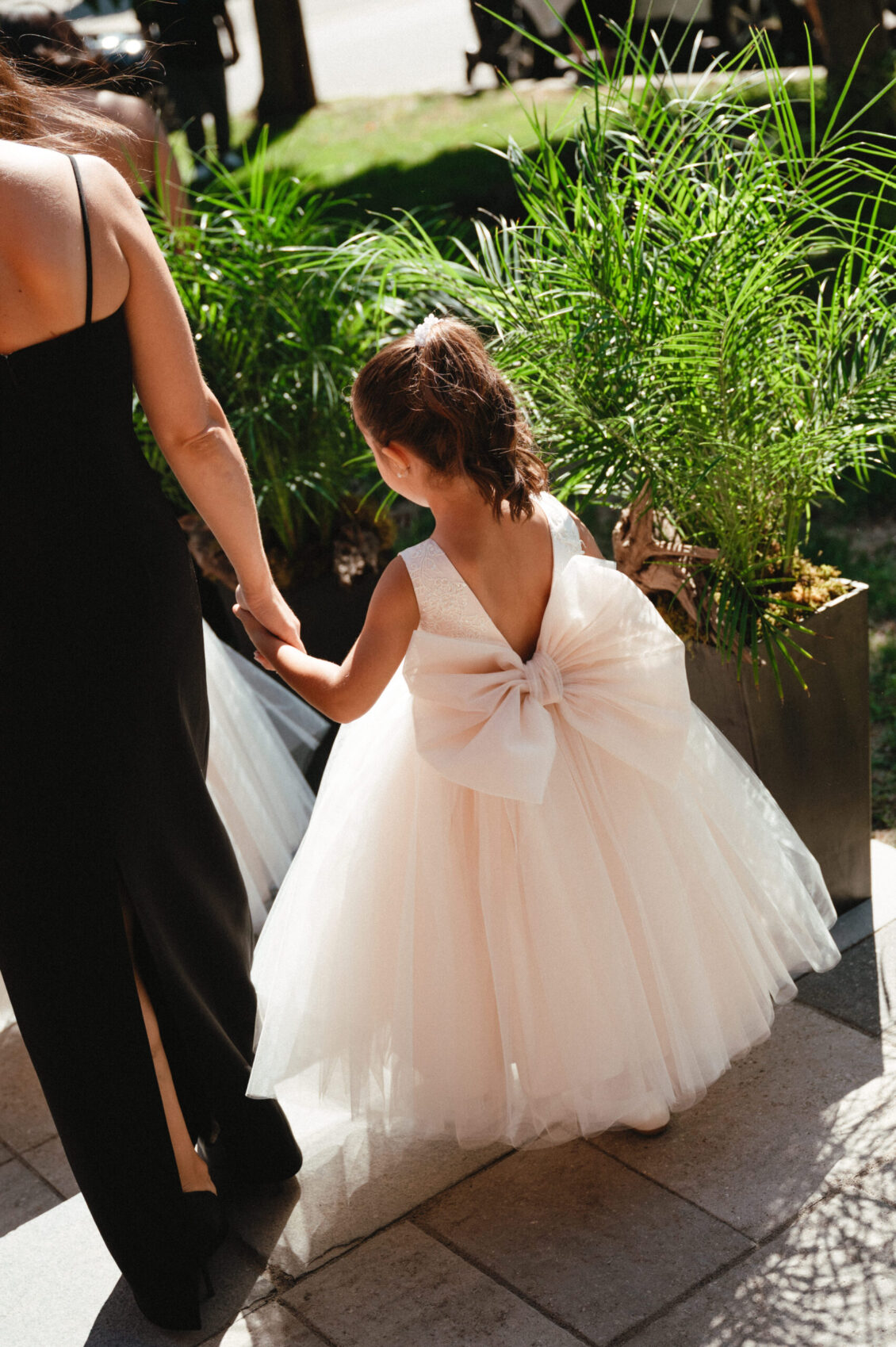 A young girl in a white dress with a large bow holds an adult’s hand while walking outside on a sunny day, surrounded by green potted plants, capturing the timeless elegance of a black-tie wedding at Hotel X Toronto.