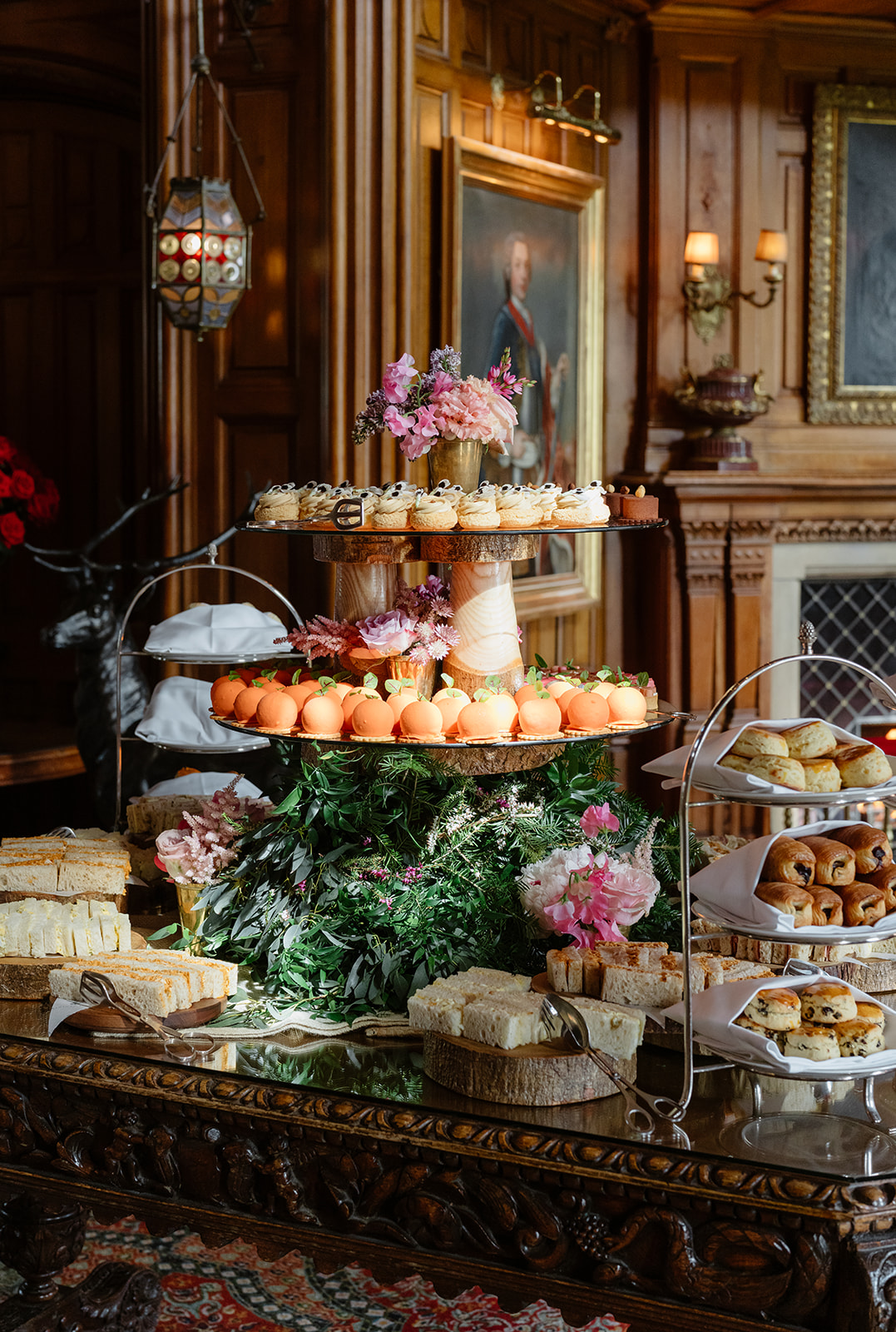 A tiered stand with cupcakes, orange macarons, flowers, and greenery, surrounded by plates of sandwiches, scones, and pastries, set in an ornate wood-paneled room with framed paintings and vintage decor—perfect for an elegant Irish castle wedding at Ashford Castle.