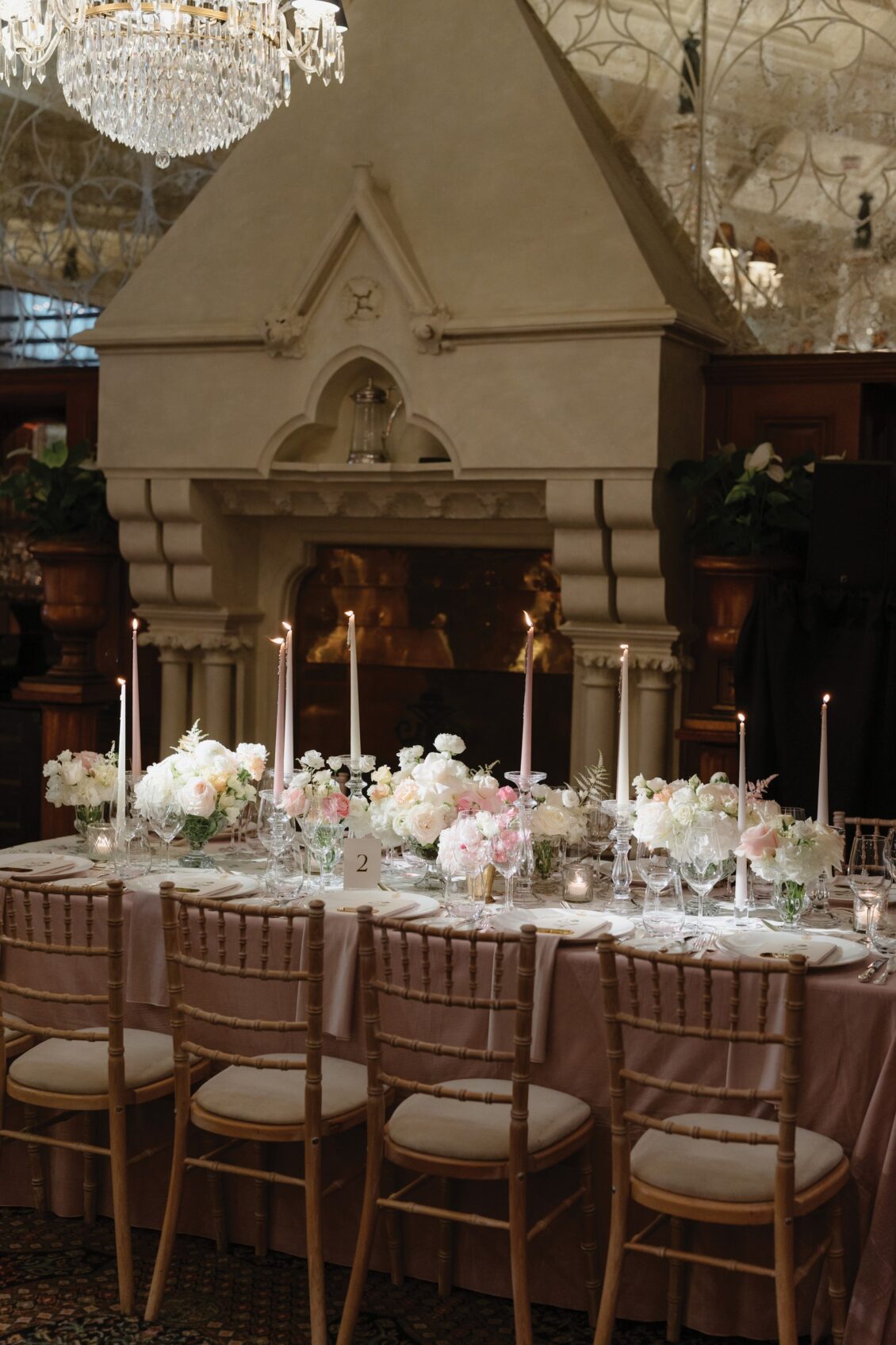 A long dining table with a pink tablecloth, elegant floral centerpieces, and tall candles set in front of a grand fireplace, surrounded by wooden chairs in a sophisticated, warmly lit room—perfectly capturing the romantic ambiance of an Irish castle wedding at Ashford Castle.