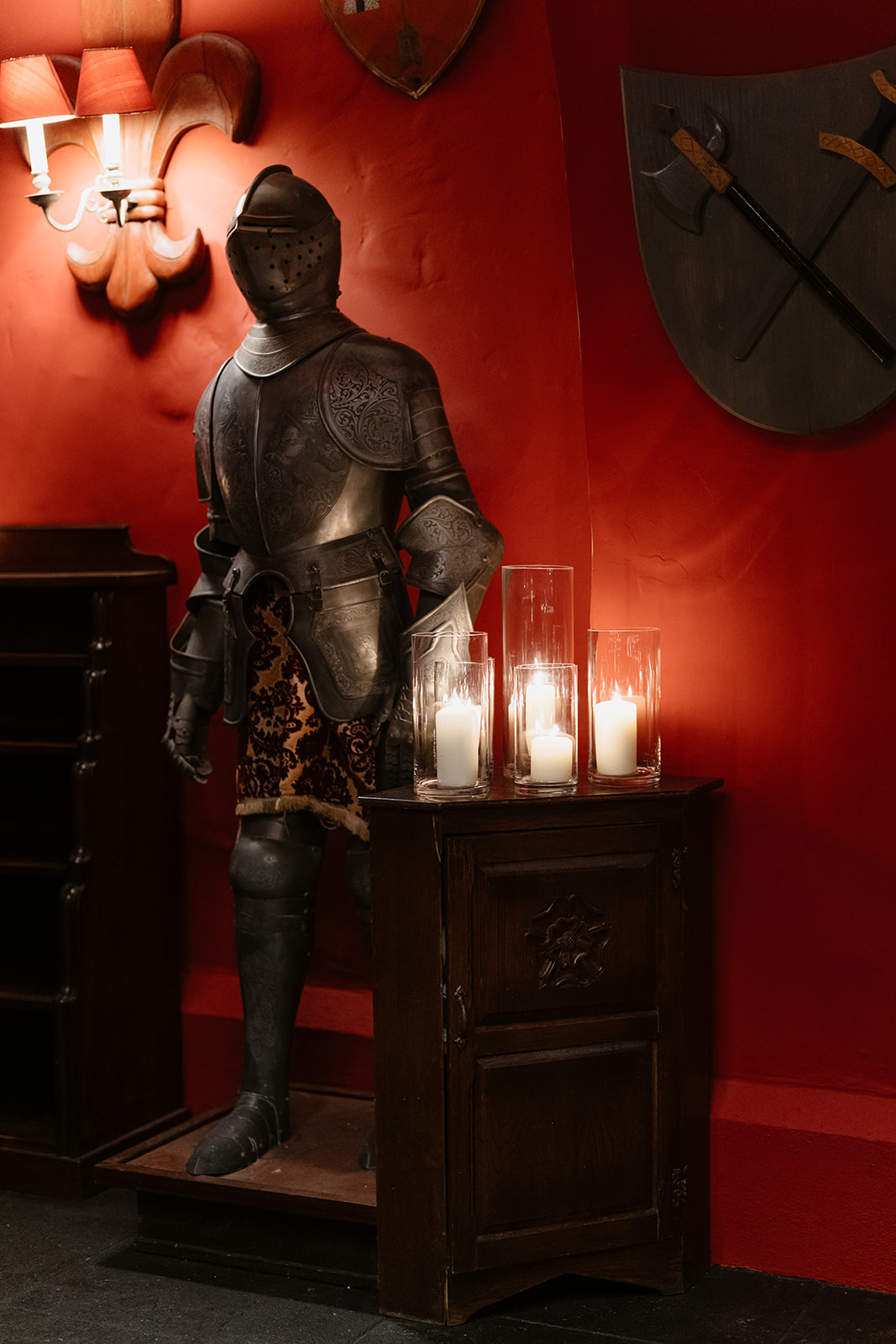 A suit of medieval armor stands beside a small wooden cabinet with three lit candles in glass holders, set against a red wall adorned with shields and a wall lamp—evoking the enchanting atmosphere of an Irish castle wedding at Ashford Castle.