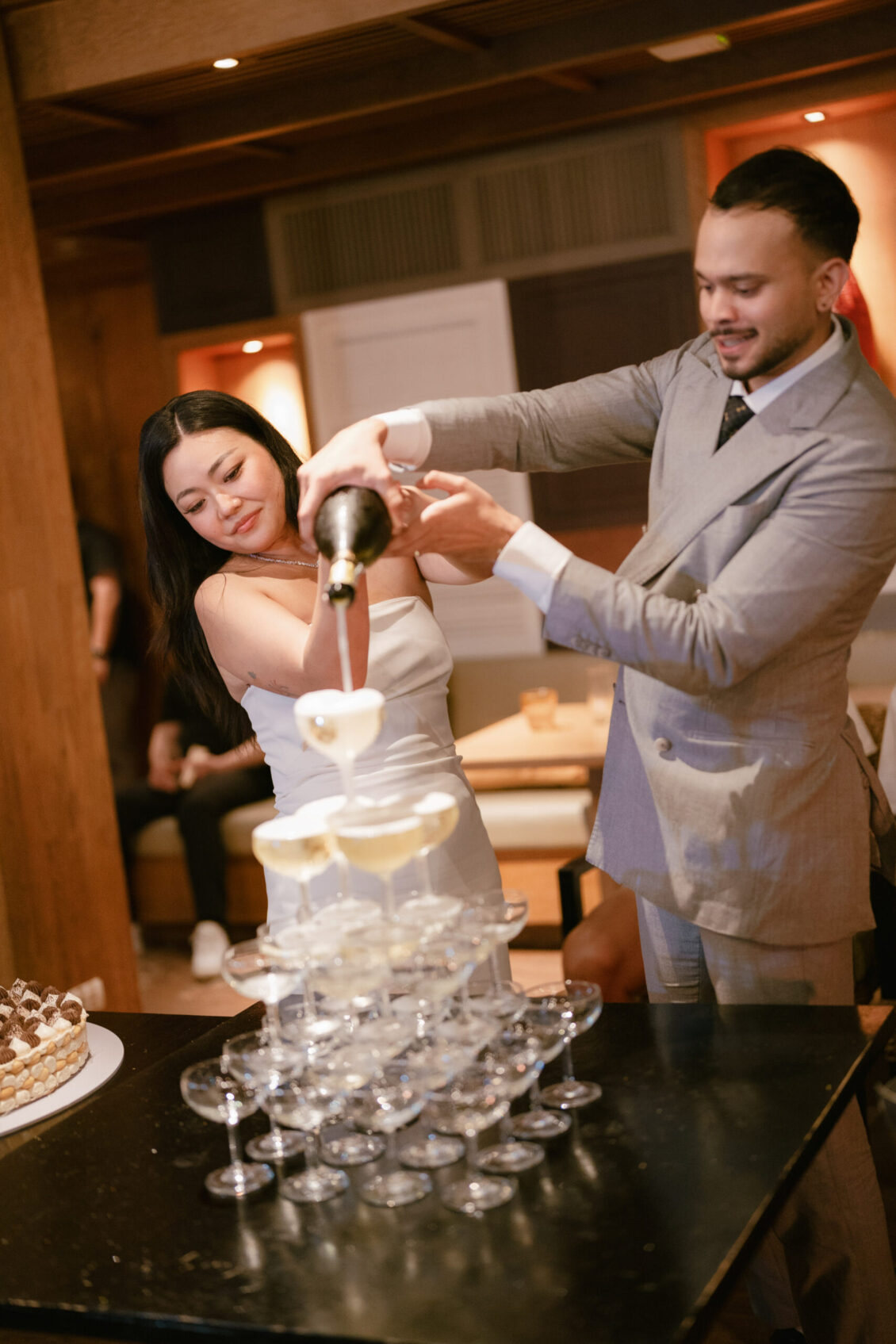 At a Renaissance Phuket wedding, a woman in a white dress and a man in a gray suit pour champagne together into a tower of stacked glasses at an indoor celebration. A cake sits on the table nearby.