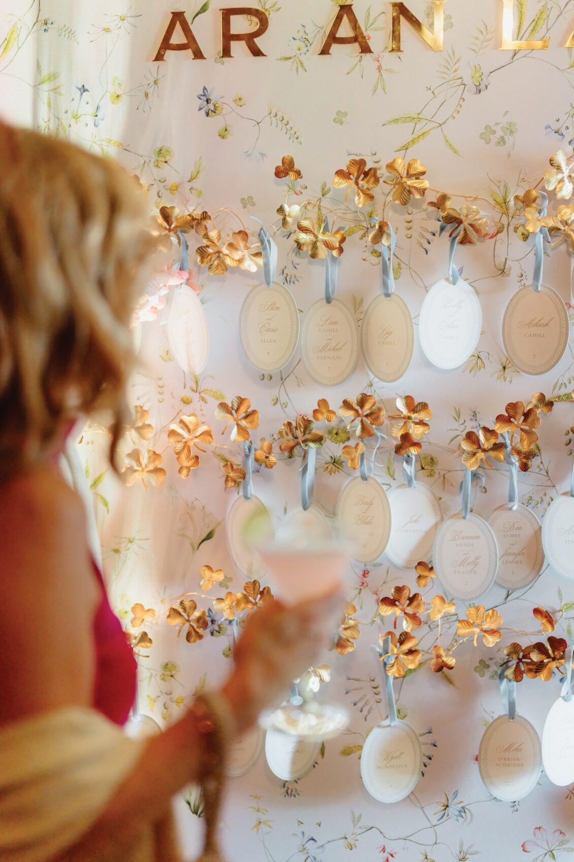 A woman holding a drink looks at a decorative wall with small framed cards and gold flowers, each card hanging on a blue ribbon—possibly an elegant seating arrangement or event display for an Irish castle wedding at Ashford Castle.