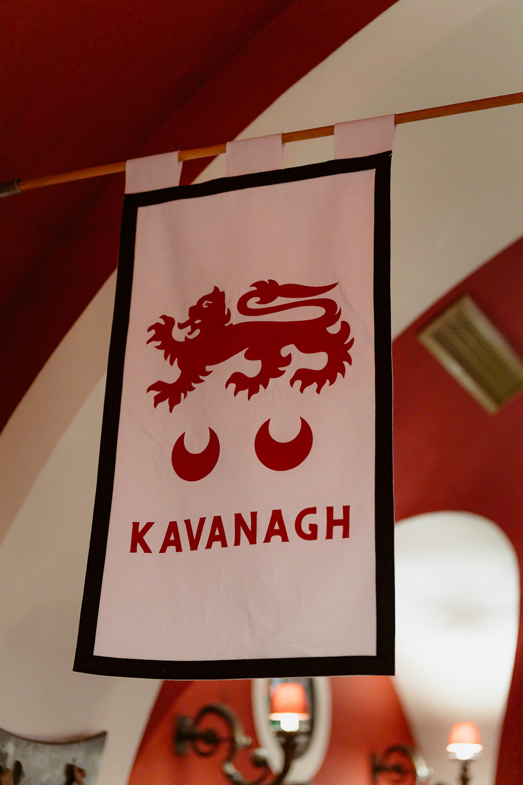 A white banner with a red lion and two red crescents above the name “Kavanagh” hangs indoors against a red and white wall background, evoking the regal charm of an Irish castle wedding at Ashford Castle.
