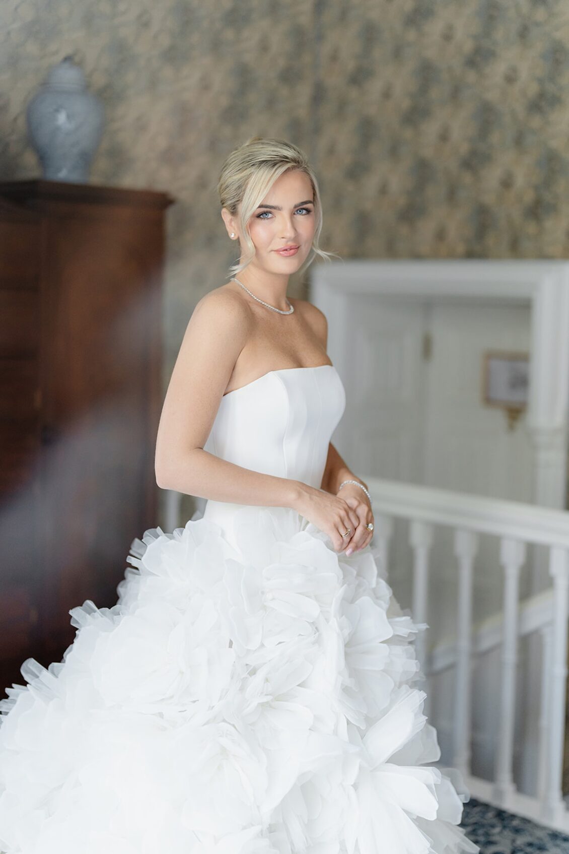 A woman in a strapless white wedding dress with a ruffled skirt stands indoors, smiling softly, with a blurred background featuring a wooden cabinet and patterned wallpaper—a serene moment from her Irish castle wedding at Ashford Castle.