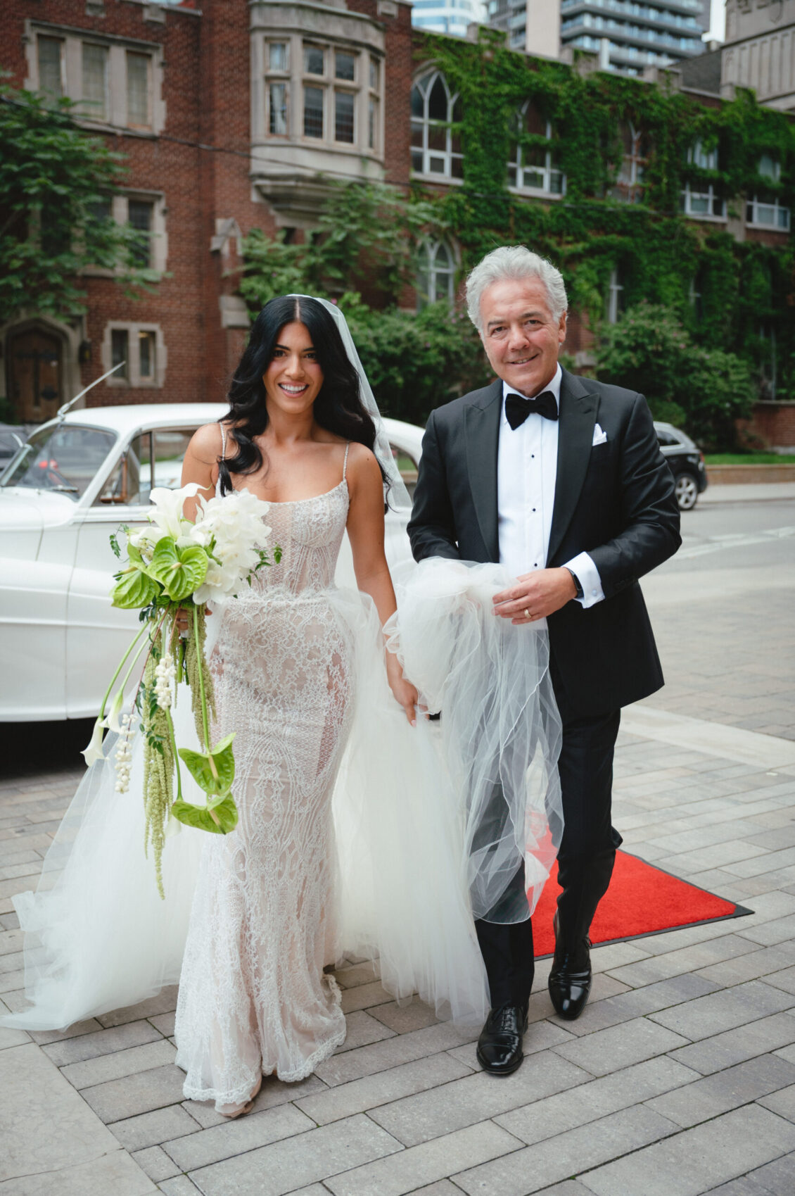 A bride in a lace wedding dress, holding a bouquet, walks with a smiling older man in a tuxedo. They are outside on a sidewalk near a vintage white car and a red carpet, in front of ivy-covered brick buildings—capturing the elegance of a black-tie wedding at Hotel X Toronto.