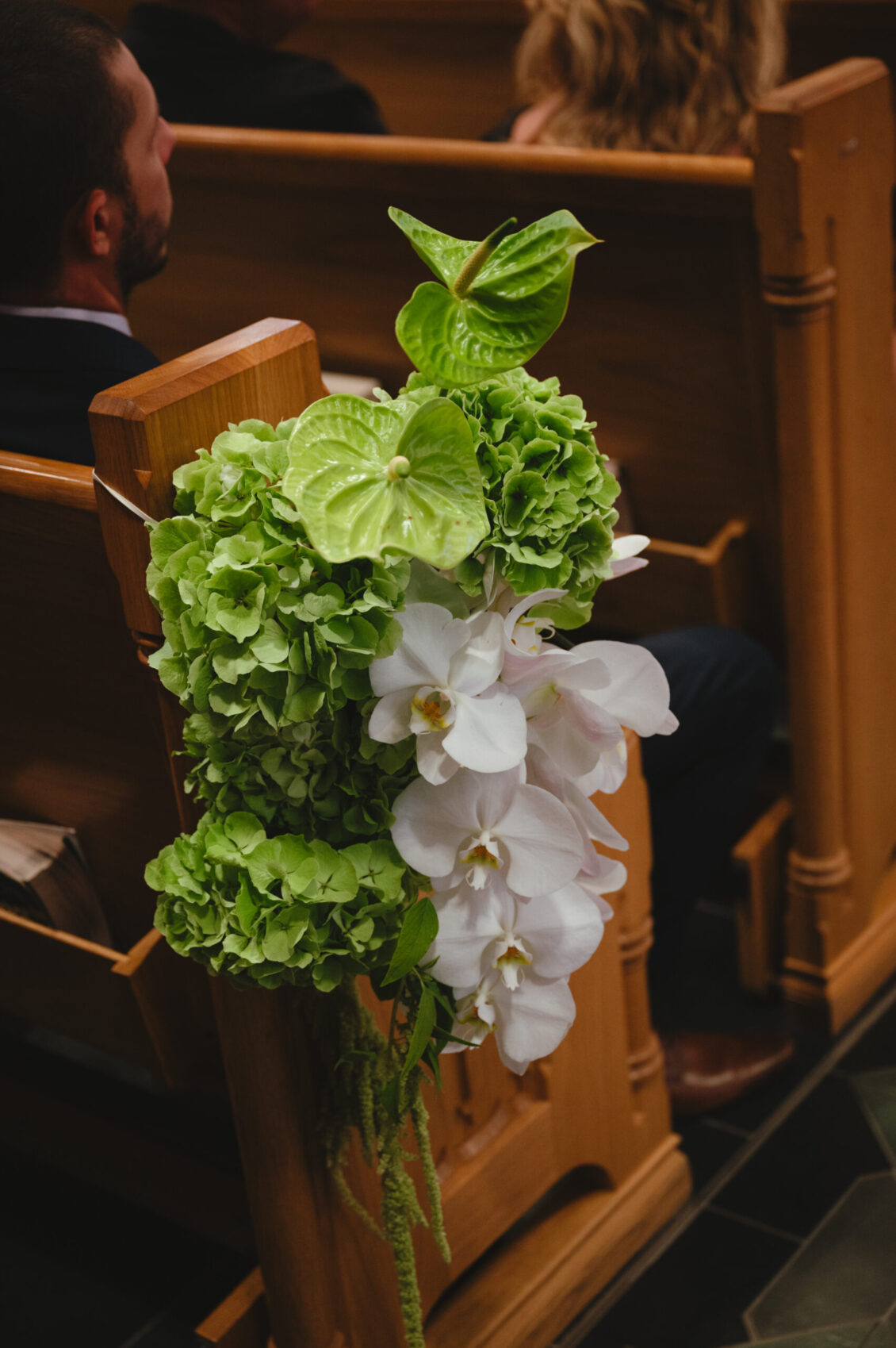 Green hydrangeas and white orchids decorate a wooden church pew at a black-tie wedding at Hotel X Toronto. People are seated in the background, slightly out of focus.