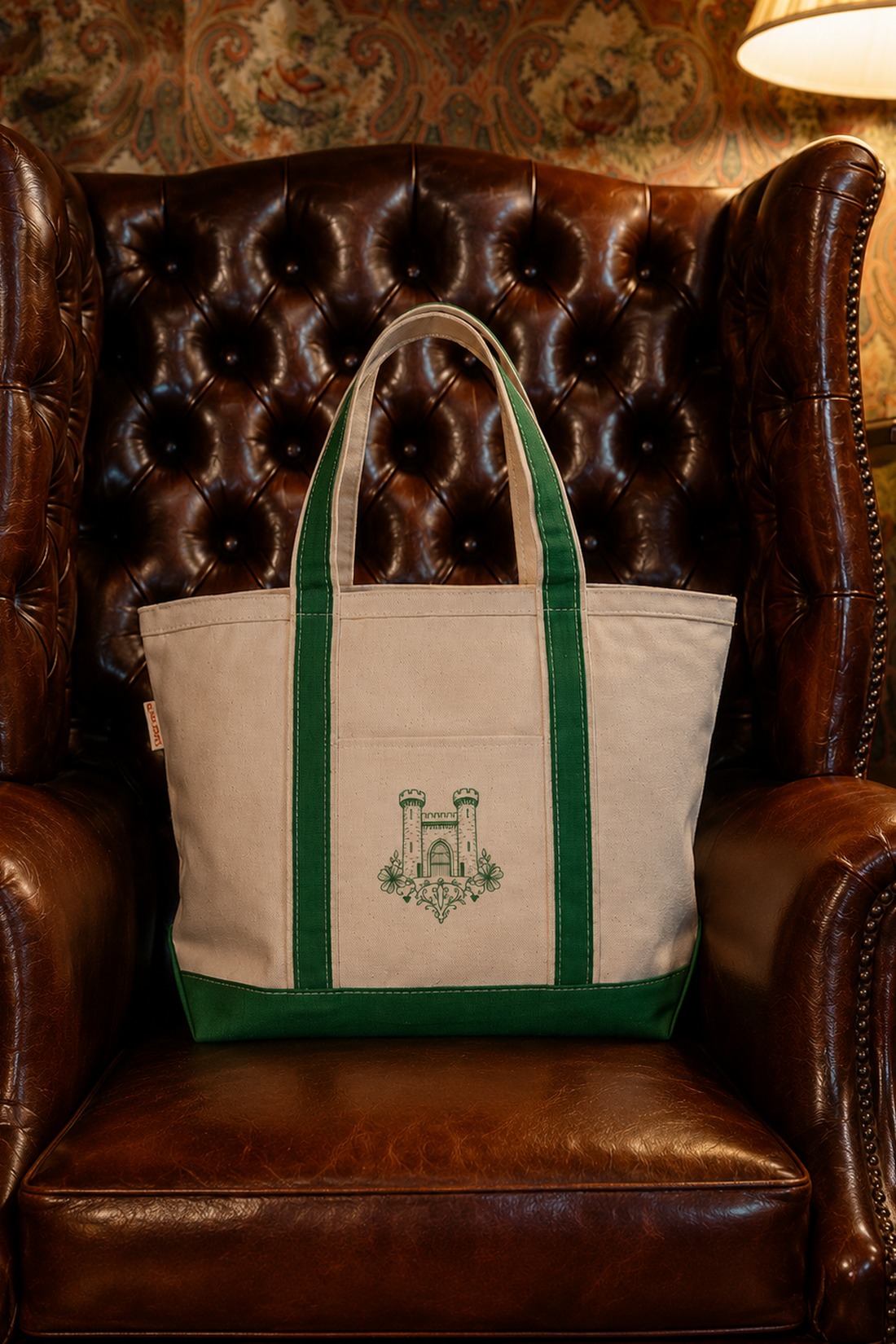 A beige tote bag with green straps and a castle emblem sits on a brown tufted leather armchair, with a lamp and patterned wallpaper in the background.