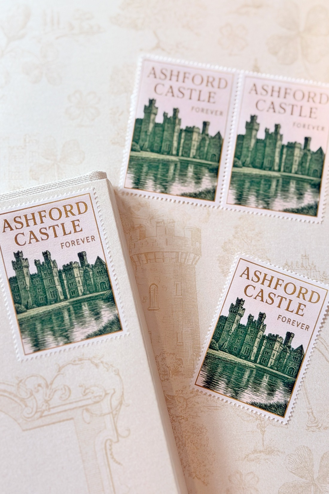 Three “Ashford Castle Forever” stamps, featuring a green illustration of a castle by water, are arranged on a cream-colored, textured background with castle-themed patterns. One stamp is attached to a box.