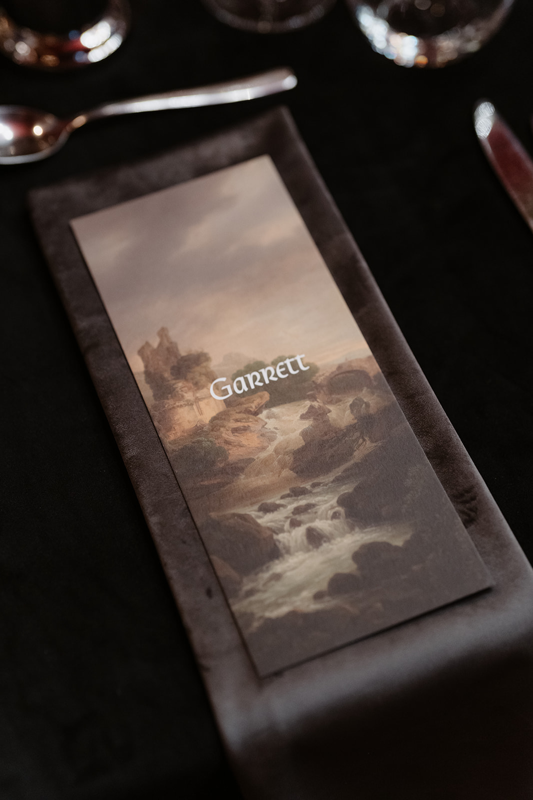 A folded menu or card with the name “Garrett” lies on a dark napkin, featuring an image of a rocky river landscape, surrounded by glassware and silverware on a black tablecloth—capturing the elegance and ambiance of an Irish castle wedding at Ashford Castle.