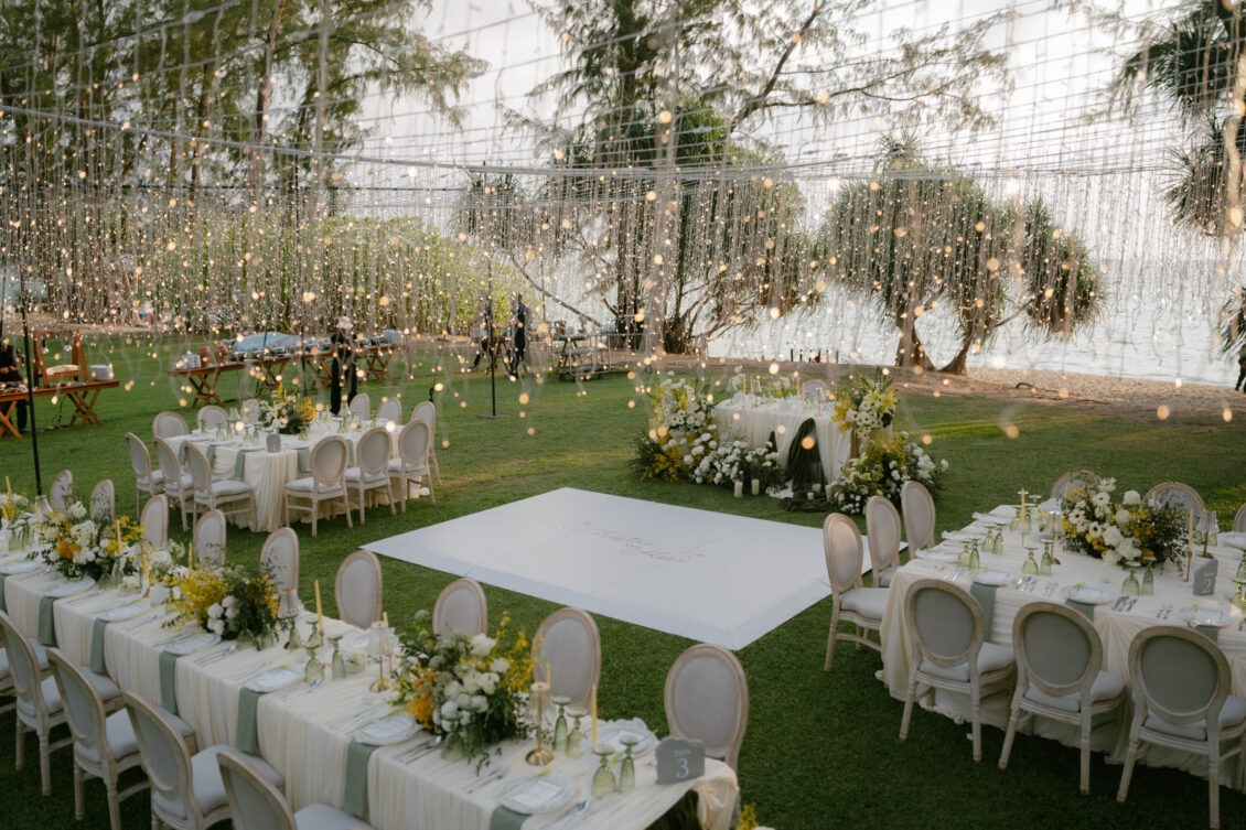 Elegant outdoor wedding setup with white tables, chairs, floral centerpieces, string lights overhead, and a central dance floor on green grass near water and trees, capturing the enchanting atmosphere of a Renaissance Phuket wedding.
