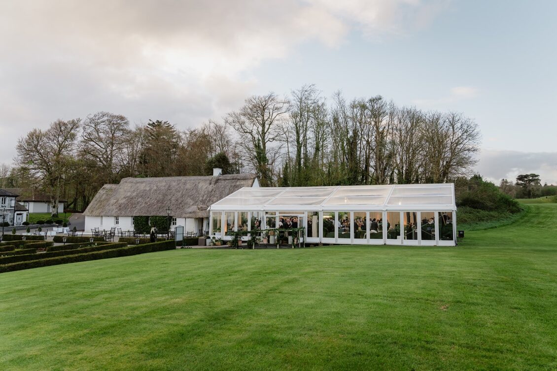 A white marquee set up on a green lawn beside a thatched cottage, surrounded by trees and hedges under a partly cloudy sky, evokes the timeless elegance of an Irish castle wedding at Ashford Castle.