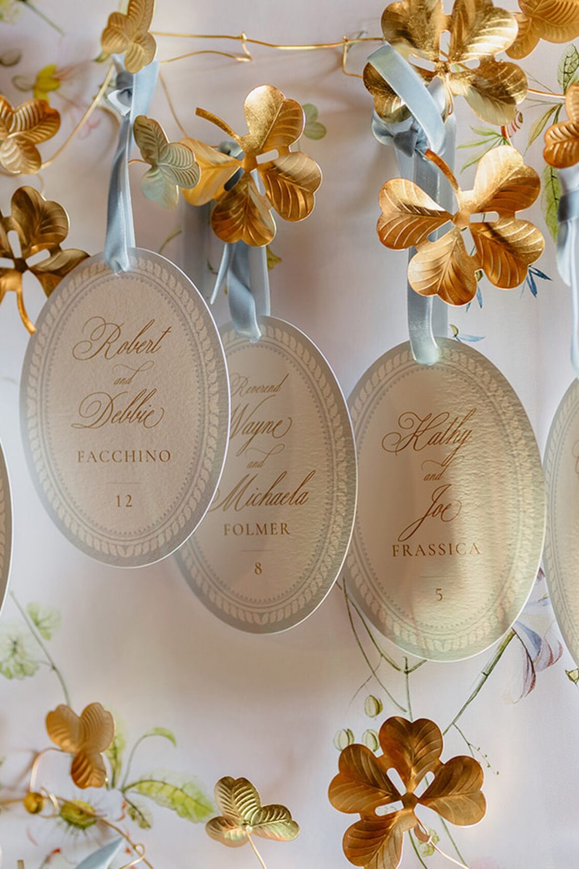 Oval name cards with gold calligraphy and blue ribbons hang among metallic gold leaves and flowers on a decorative wall, perfectly setting the scene for an opulent Irish castle wedding at Ashford Castle. The cards display names and table numbers for a formal event or wedding.