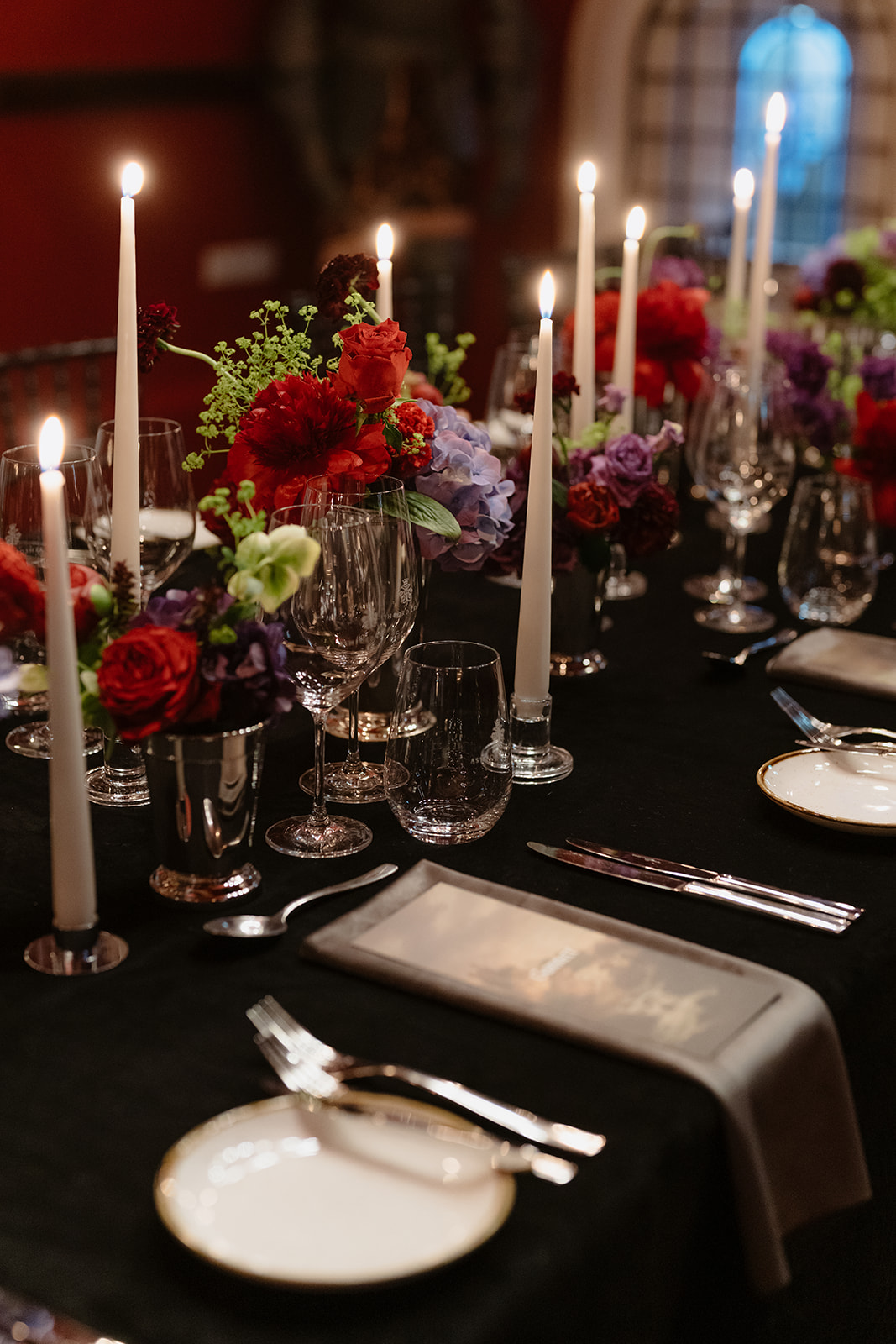 Elegant dinner table set with white candles, vibrant floral arrangements, glassware, plates, and silverware on a black tablecloth. Warm, ambient lighting creates a cozy atmosphere reminiscent of an Irish castle wedding at Ashford Castle.