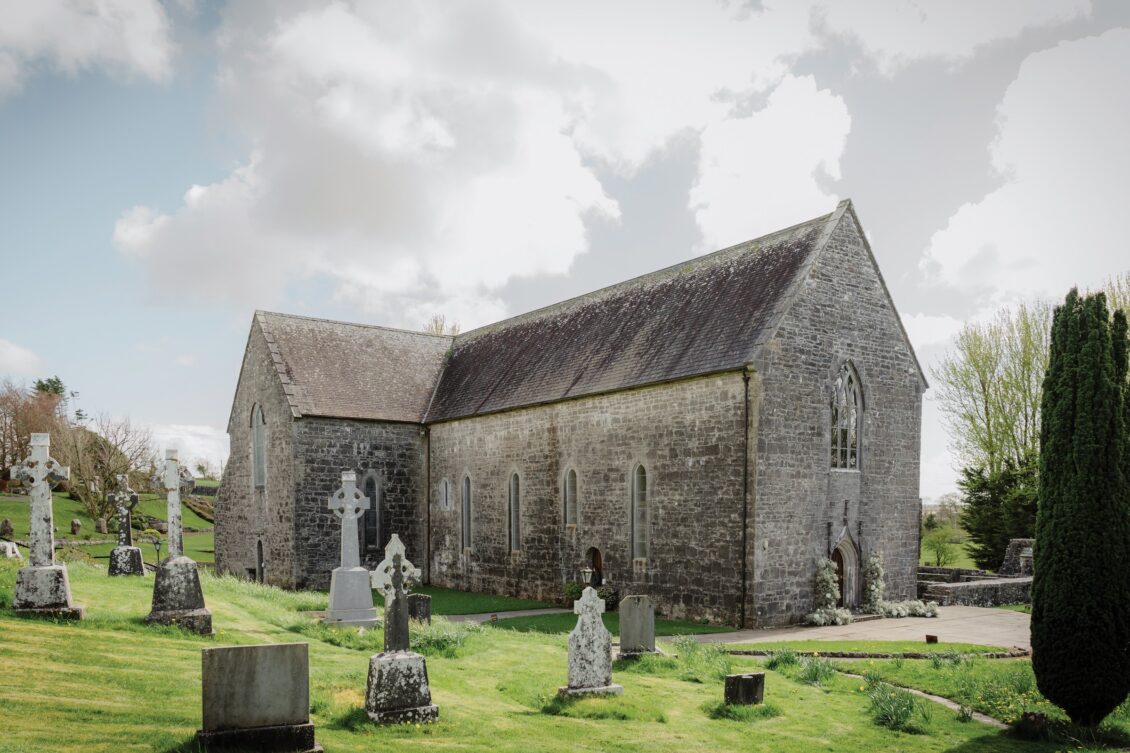 An old stone church stands among gravestones on a grassy hill under a partly cloudy sky, surrounded by trees and greenery, evoking the timeless romance of an Irish castle wedding at Ashford Castle.