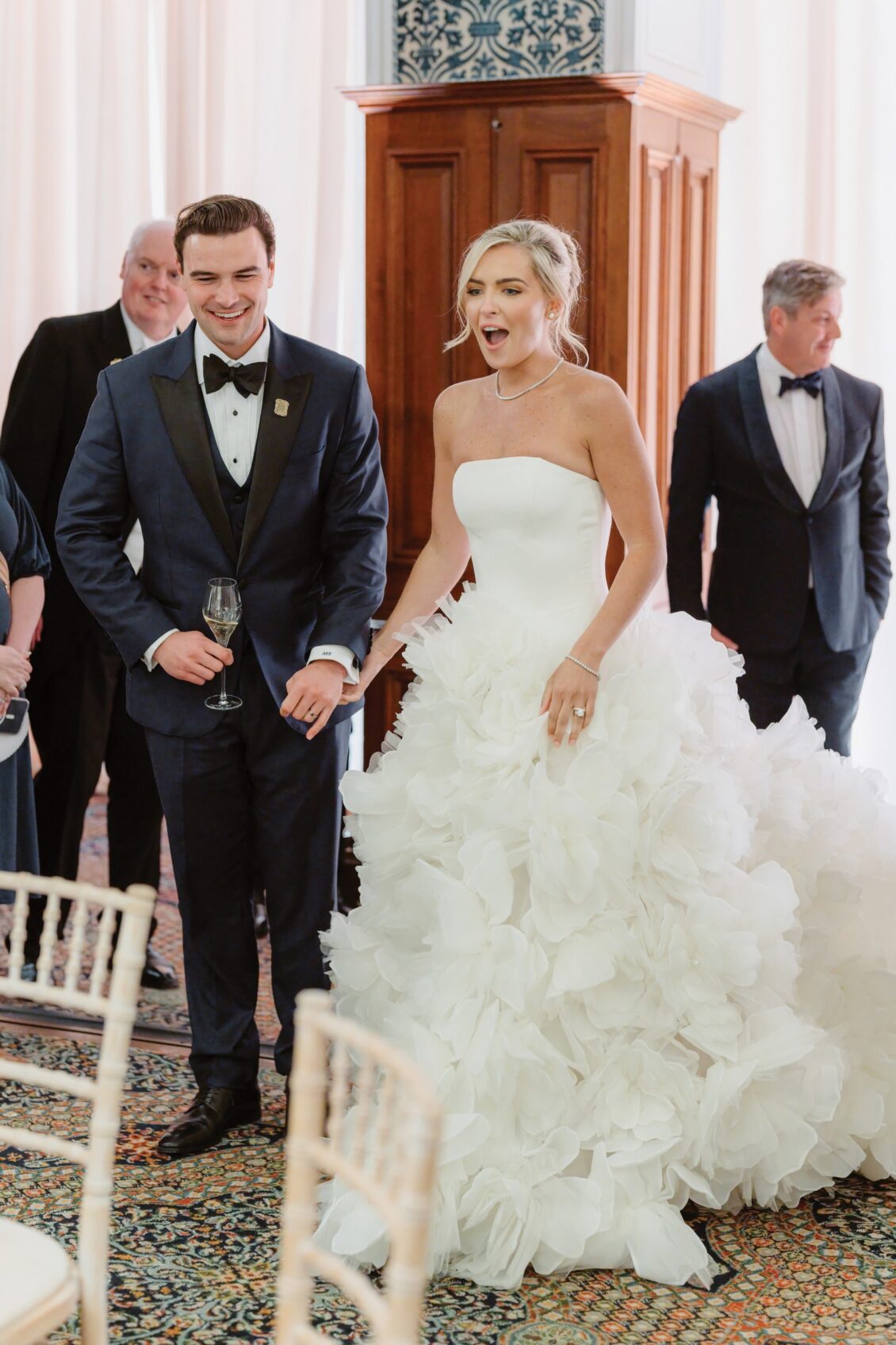 A bride in a strapless, ruffled white gown stands beside a groom in a dark tuxedo, both smiling. Guests in formal attire are visible in the background, and the elegant and festive setting evokes the timeless romance of an Irish castle wedding at Ashford Castle.