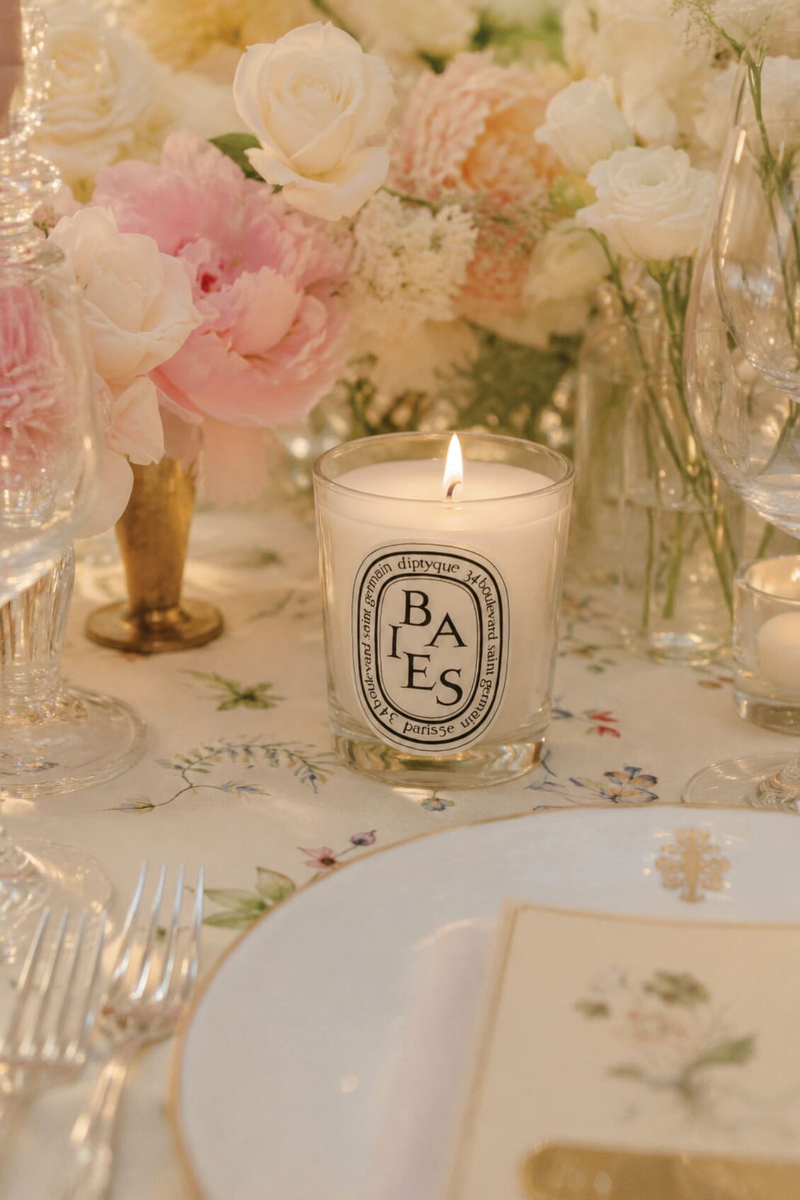A lit Diptyque Baies candle sits on a floral tablecloth, surrounded by elegant glassware, white and pink flowers, and a white plate with a floral card—capturing the romantic ambiance of an Irish castle wedding at Ashford Castle.