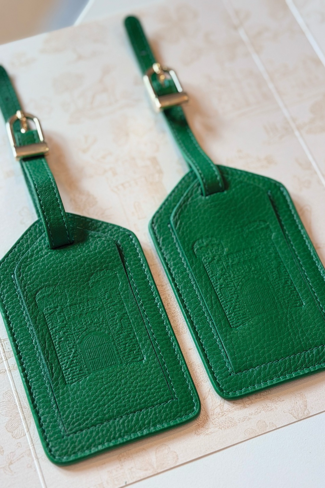 Two green leather luggage tags with embossed architectural designs and silver buckles are placed side by side on a patterned surface.