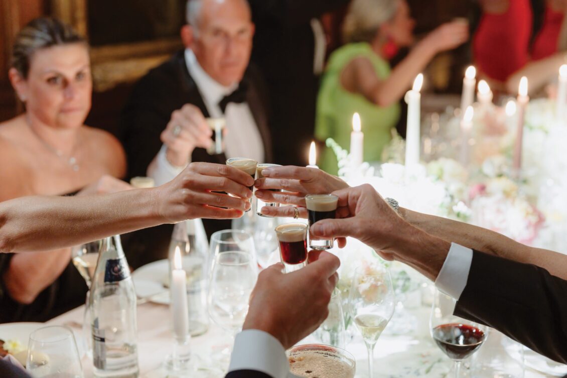 People in formal attire clink shot glasses at a candlelit dinner table, surrounded by glasses, bottles, and flowers, creating a celebratory atmosphere reminiscent of an Irish castle wedding at Ashford Castle.