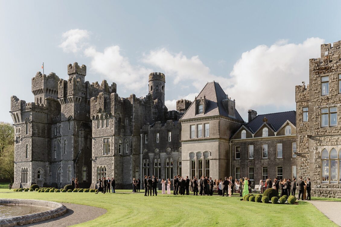 A large group of people in formal attire gathers on the lawn outside the grand Ashford Castle, its majestic towers and turrets standing tall under a partly cloudy sky, capturing the elegance of an Irish castle wedding.