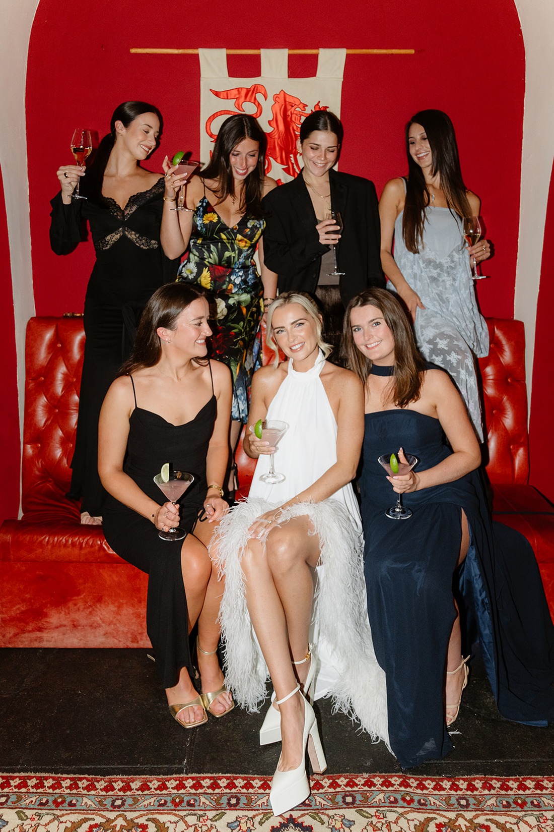 Seven women dressed formally pose with drinks on a red couch against a red wall with a decorative banner, capturing the elegant atmosphere of an Irish castle wedding at Ashford Castle. Some are seated while others stand behind, all smiling and looking at each other, radiating joy and celebration.