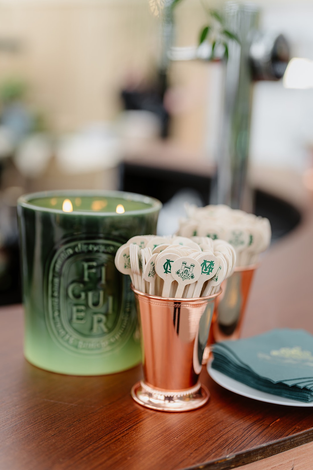 A green candle labeled “Figuier” sits beside two copper cups holding decorative stir sticks and a stack of green napkins on a wooden surface, evoking the elegant charm of an Irish castle wedding at Ashford Castle.