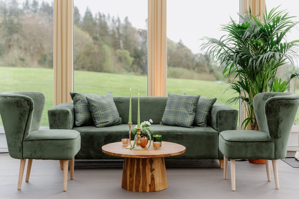 A cozy living room reminiscent of an Irish castle wedding at Ashford Castle, featuring a green velvet sofa, plaid pillows, two matching chairs, a wooden coffee table adorned with candles and decor, and a tall potted plant near large windows overlooking a grassy landscape.