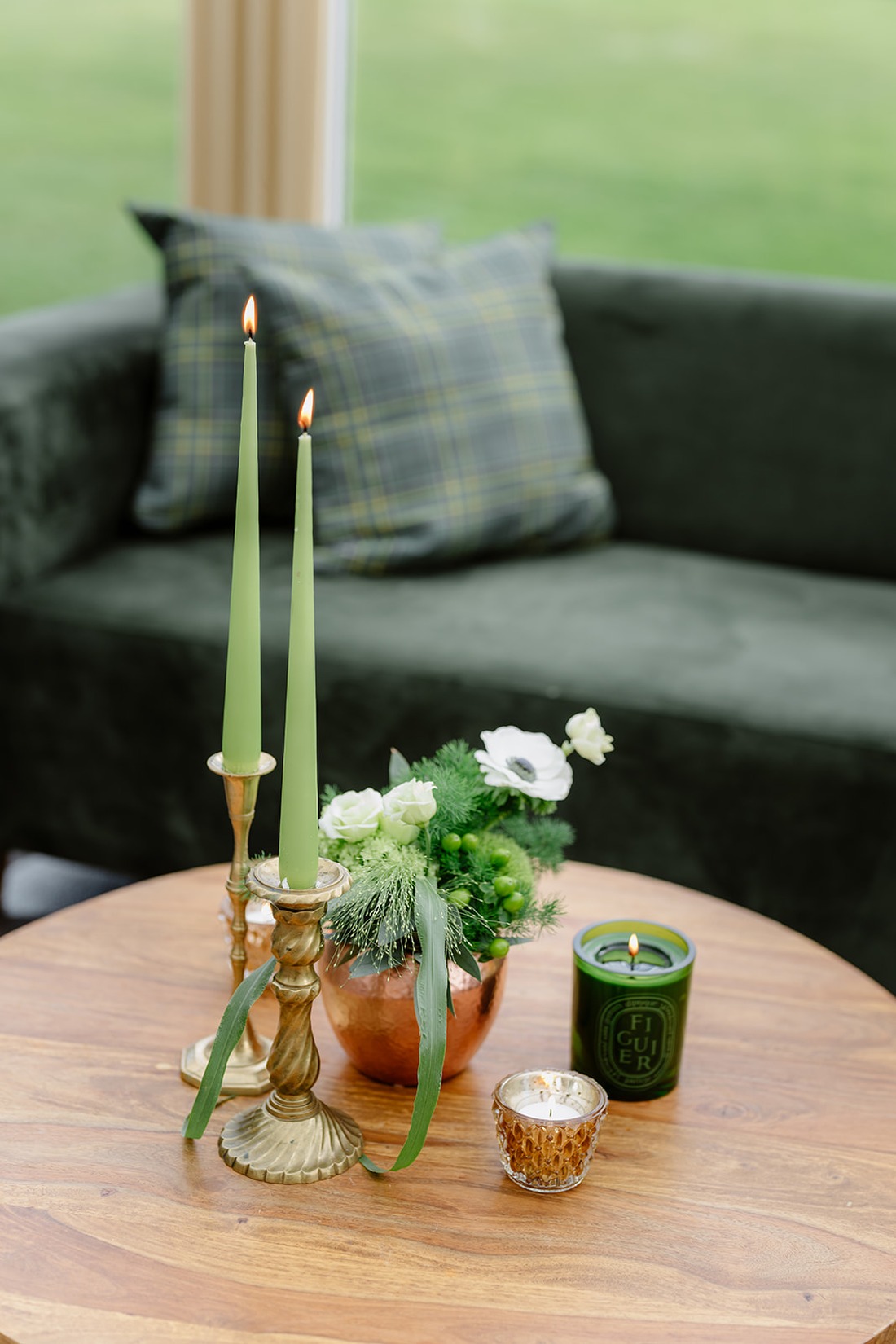 Two green taper candles in brass holders, a small floral arrangement, and two candles in glass containers sit on a round wooden table; in the background, a green velvet couch with plaid pillow evokes the timeless elegance of an Irish castle wedding at Ashford Castle.