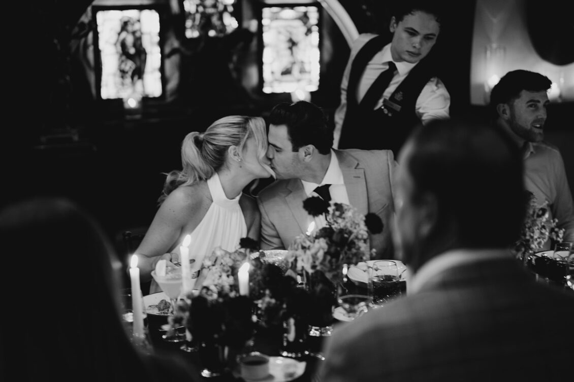 A couple kisses at a candlelit dinner table with flowers, surrounded by guests, during an Irish castle wedding at Ashford Castle. A server stands behind them. The scene is in black and white, with stained glass windows in the background.