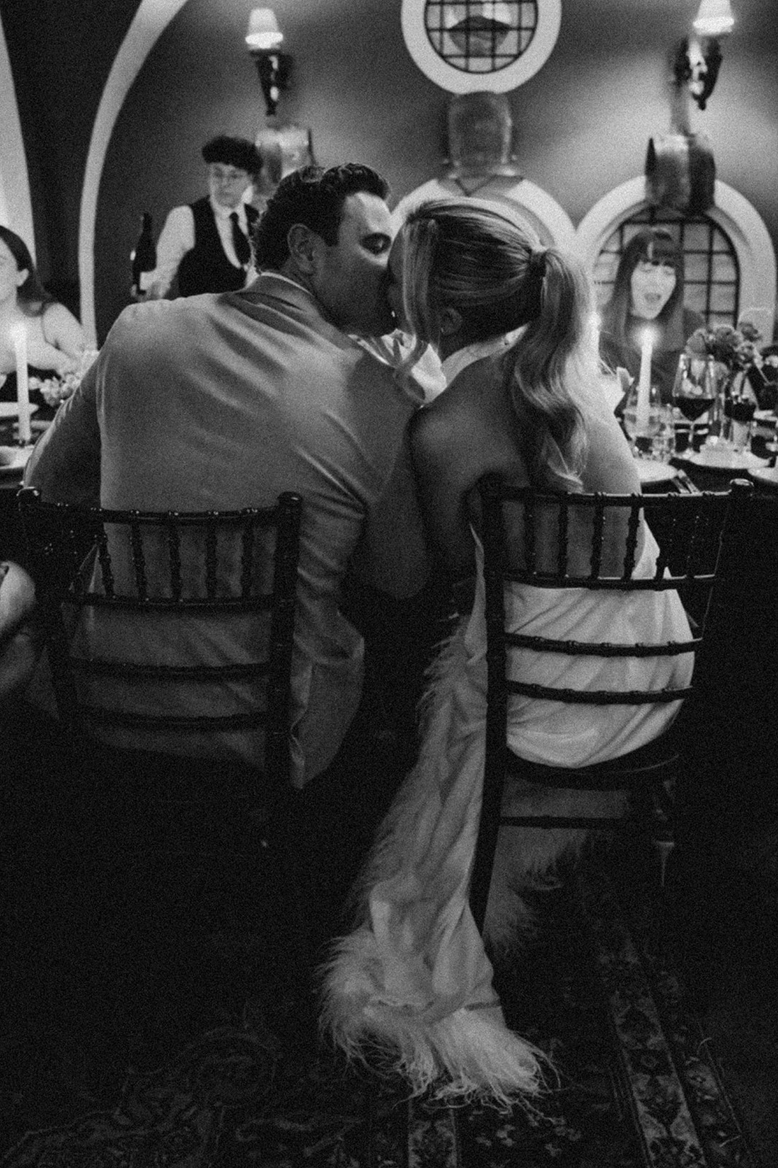 A couple sits closely together, sharing a kiss at a candlelit dinner table with other guests in a warmly decorated, intimate room during an elegant Irish castle wedding at Ashford Castle. The photo is in black and white.