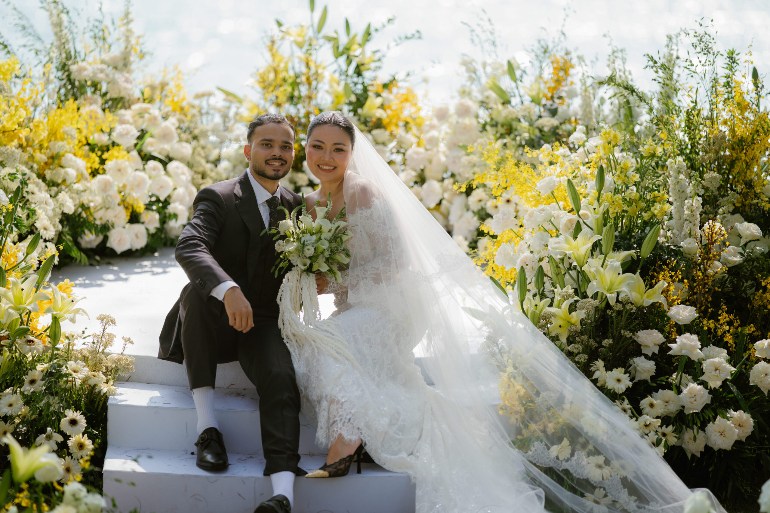 A bride and groom sit on white steps surrounded by lush yellow and white flowers, capturing the essence of a Renaissance Phuket wedding. The bride holds a bouquet and wears a lace dress with a long veil; both are smiling at the camera.