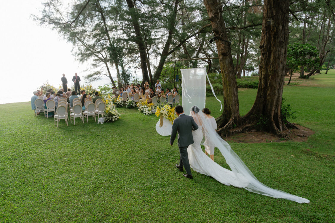 A bride and groom walk down the aisle outdoors towards seated guests and a wedding altar, surrounded by trees and greenery, capturing the enchanting ambiance of a Renaissance Phuket wedding.