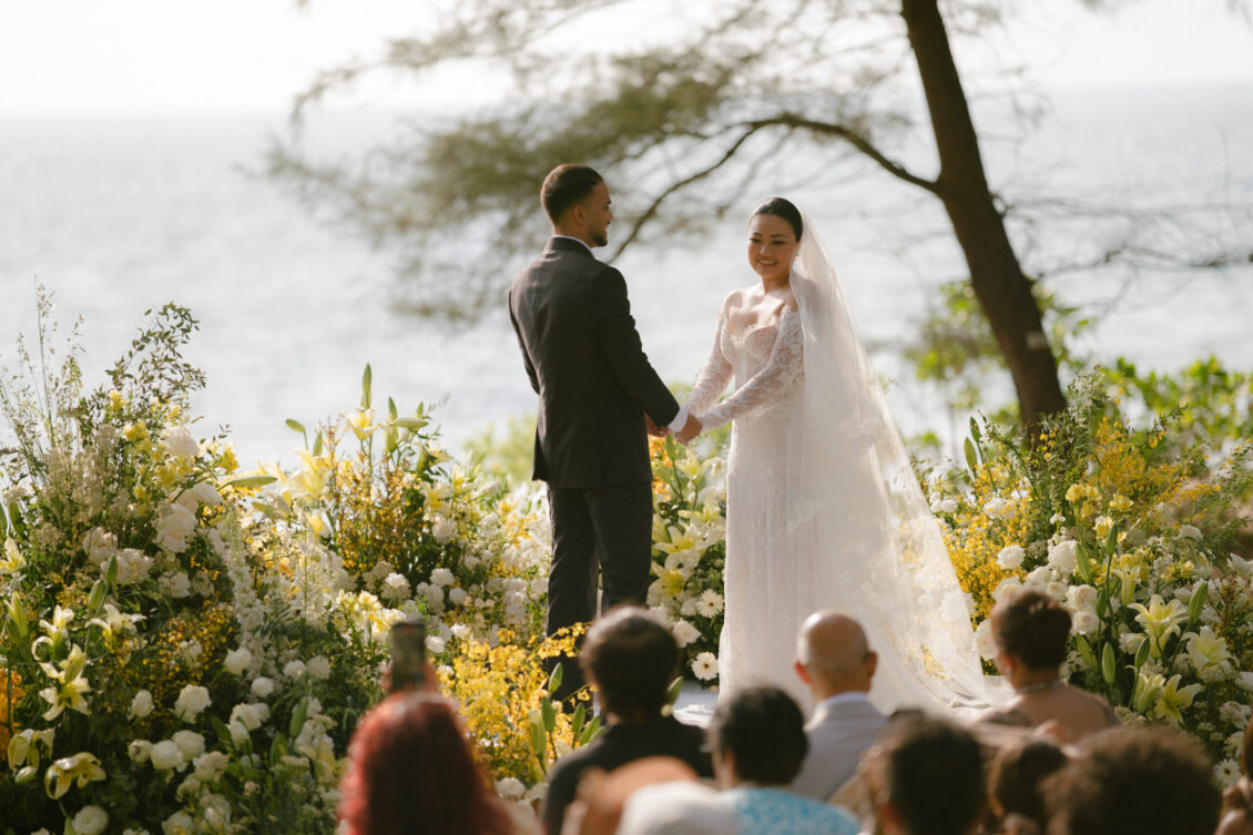 A bride and groom stand holding hands at a Renaissance Phuket wedding ceremony, surrounded by flowers, with guests seated and the ocean visible in the background.