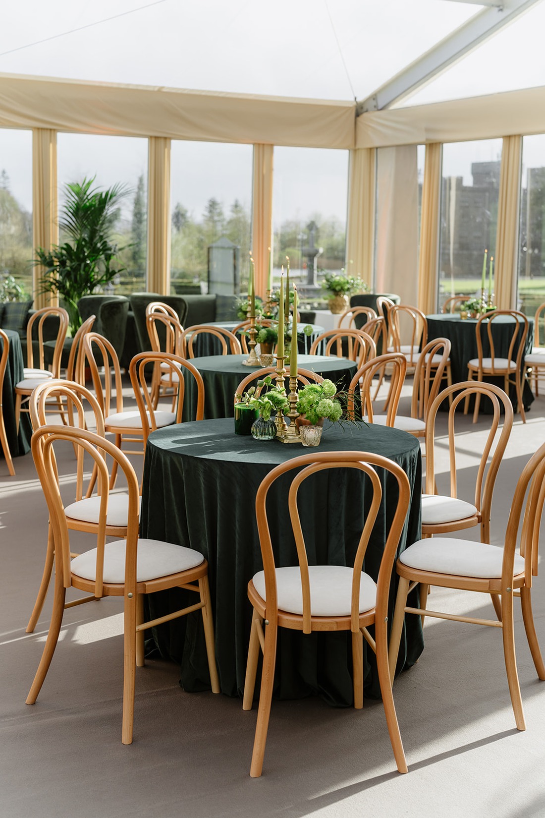 Elegant round tables with dark green tablecloths and wooden chairs are arranged in a bright, spacious room with large windows, evoking the timeless charm of an Irish castle wedding at Ashford Castle. Each table features green floral centerpieces and tall green candles, creating a refined atmosphere reminiscent of regal celebrations.