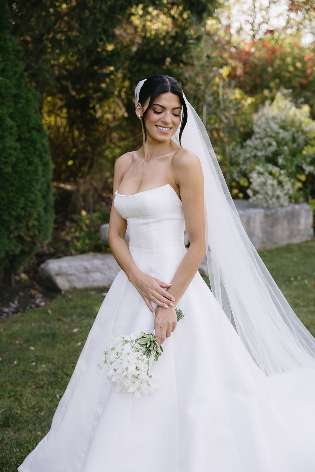A smiling bride named Erica in a strapless white wedding dress and long veil stands outdoors, holding a small bouquet of white flowers, surrounded by greenery and sunlight.