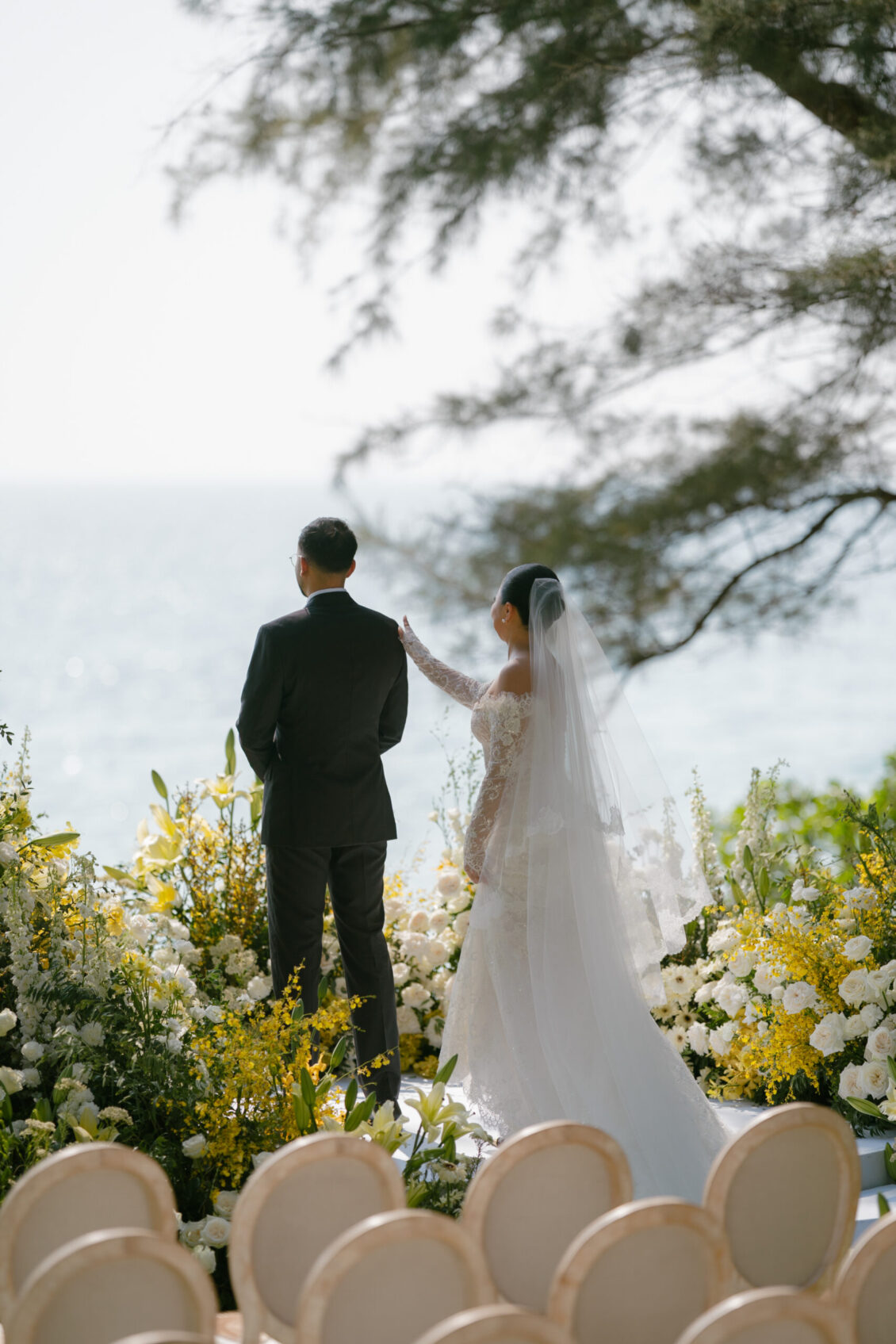 A bride in a white gown and veil gently touches the groom’s shoulder as they stand outdoors by the sea, surrounded by yellow and white flowers and empty chairs, capturing the elegance of a Renaissance Phuket wedding.
