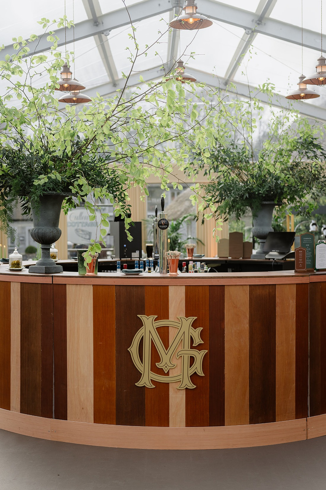 A circular wooden bar with a gold “MF” monogram, topped with bottles, glasses, and lush green floral arrangements, set under a glass ceiling with hanging lights—perfectly evoking the elegance of an Irish castle wedding at Ashford Castle.