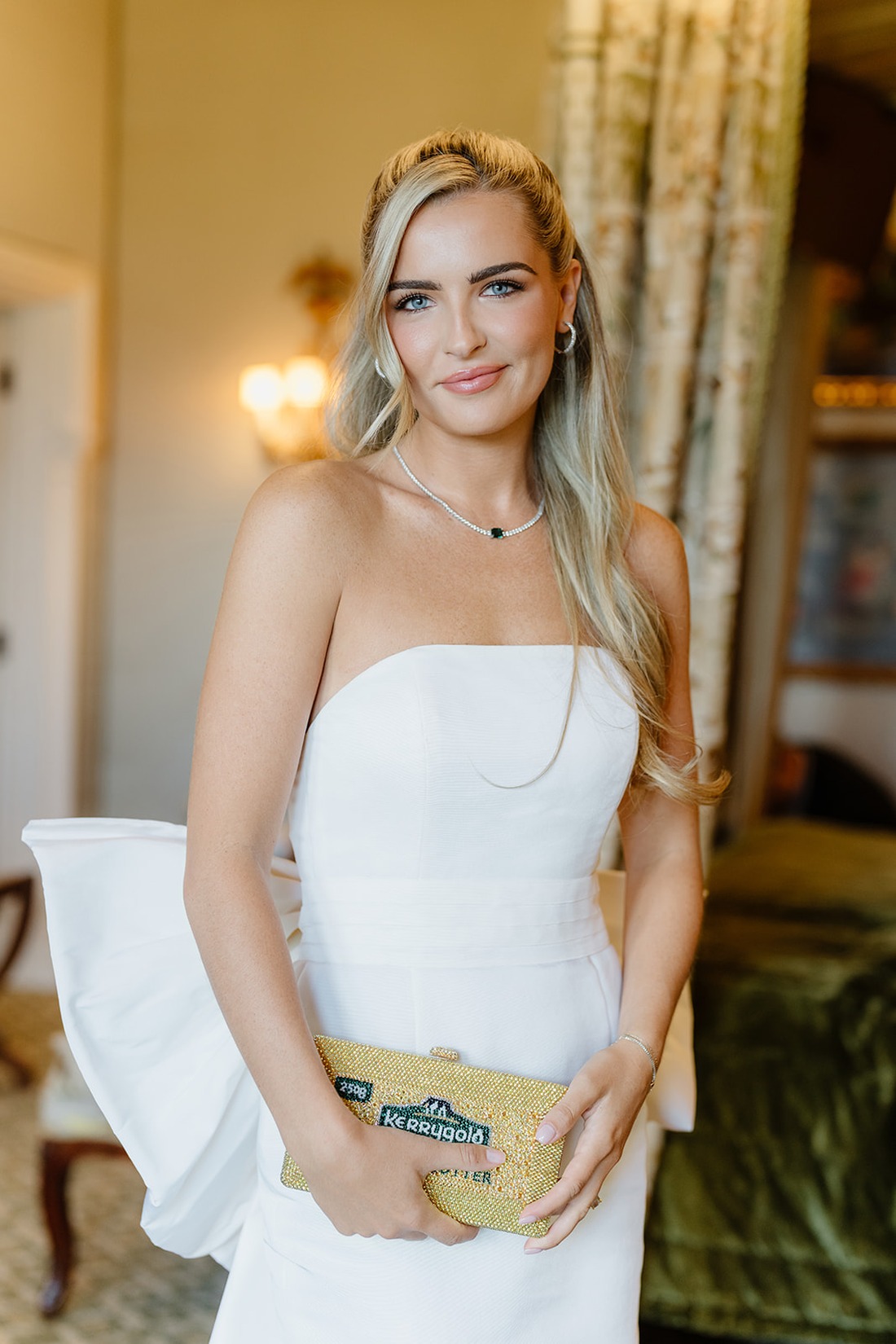A woman in a strapless white dress stands indoors at an Irish castle wedding at Ashford Castle, holding a small gold clutch, her long blonde hair styled to one side and a gentle smile on her face.