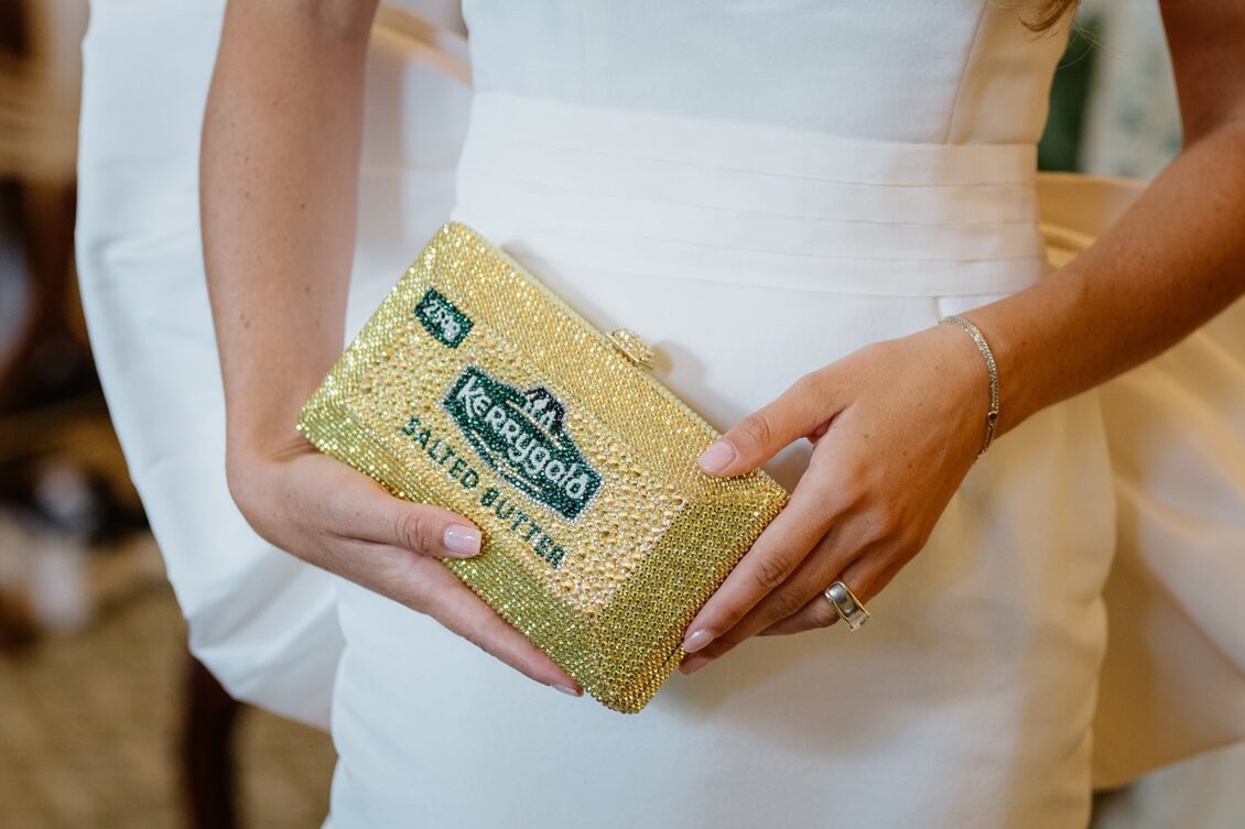 A person in a white dress holds a sparkling gold clutch designed to look like a stick of salted butter, perfect for an Irish castle wedding at Ashford Castle. The clutch features a decorative label, while the person wears a silver ring and bracelet.