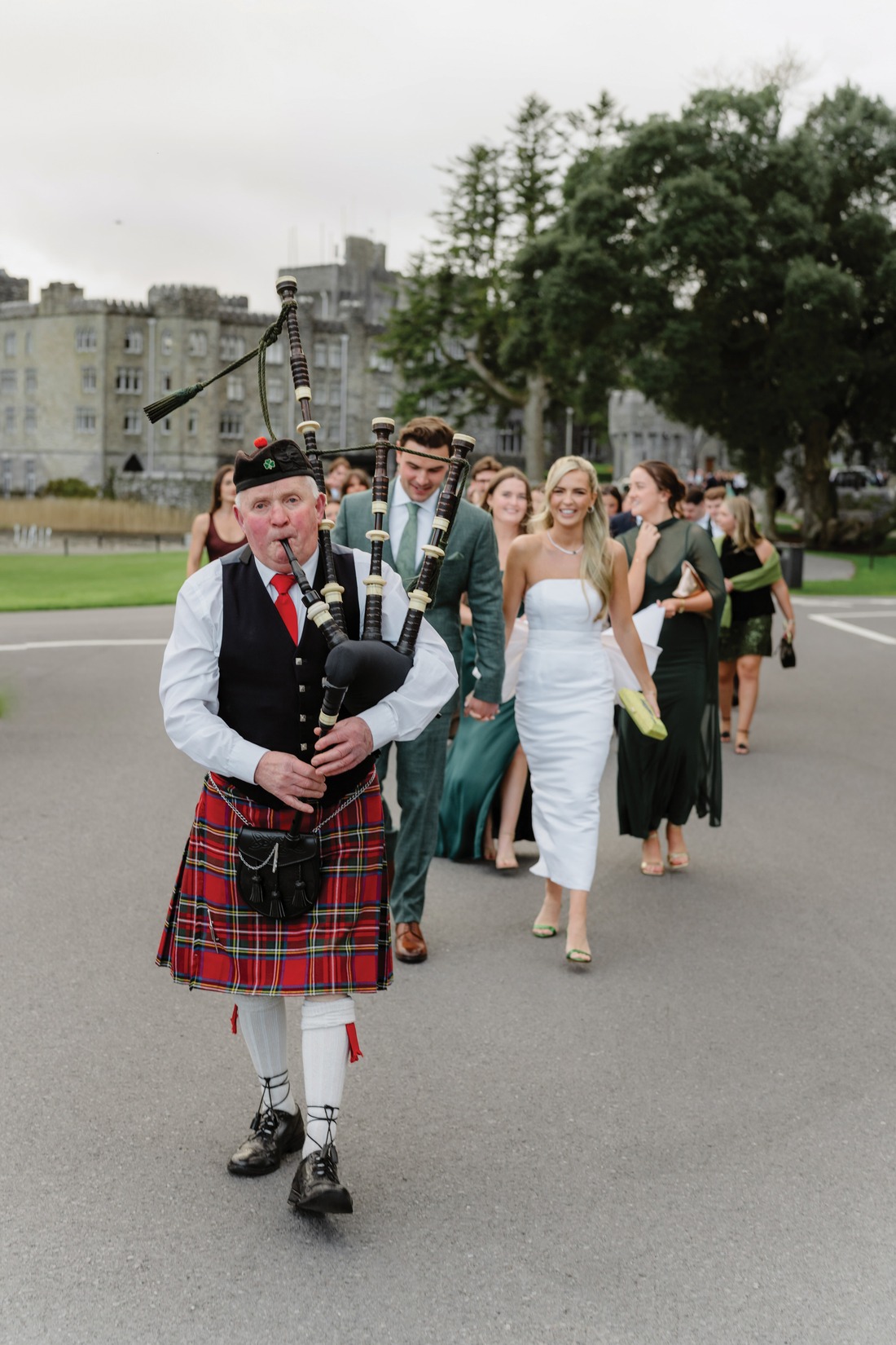 A bagpiper in a red tartan kilt leads a group of well-dressed people walking outdoors, with the majestic Ashford Castle and green trees in the background, capturing the elegance of an Irish castle wedding.